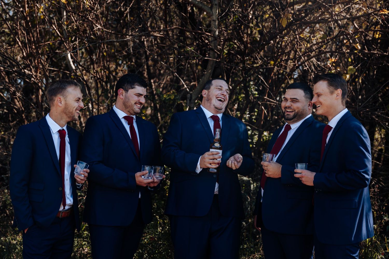 Five groomsmen in navy suits and red ties laughing and holding drinks outdoors at a wedding celebration.