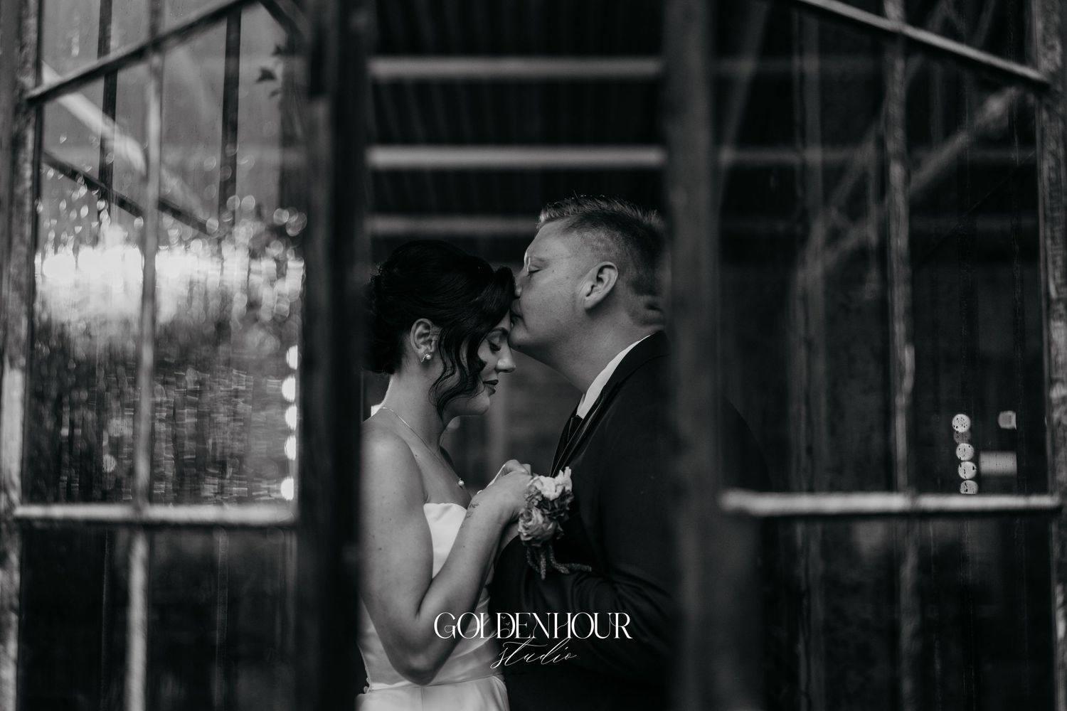 Black and white romantic photo of bride and groom sharing a kiss framed by industrial glass windows.