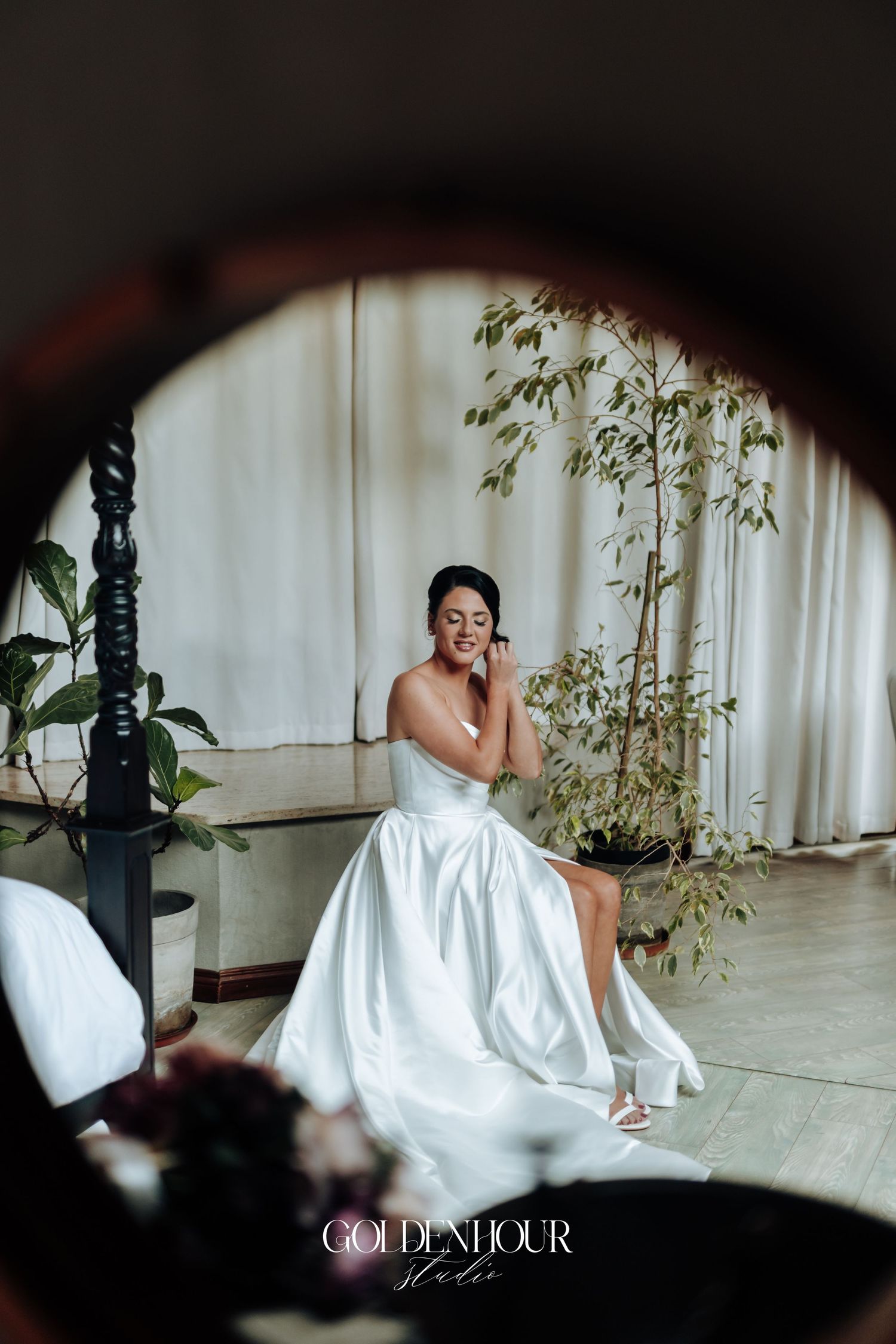 Bride in strapless white satin gown sits smiling, reflected in a round mirror during wedding preparations.
