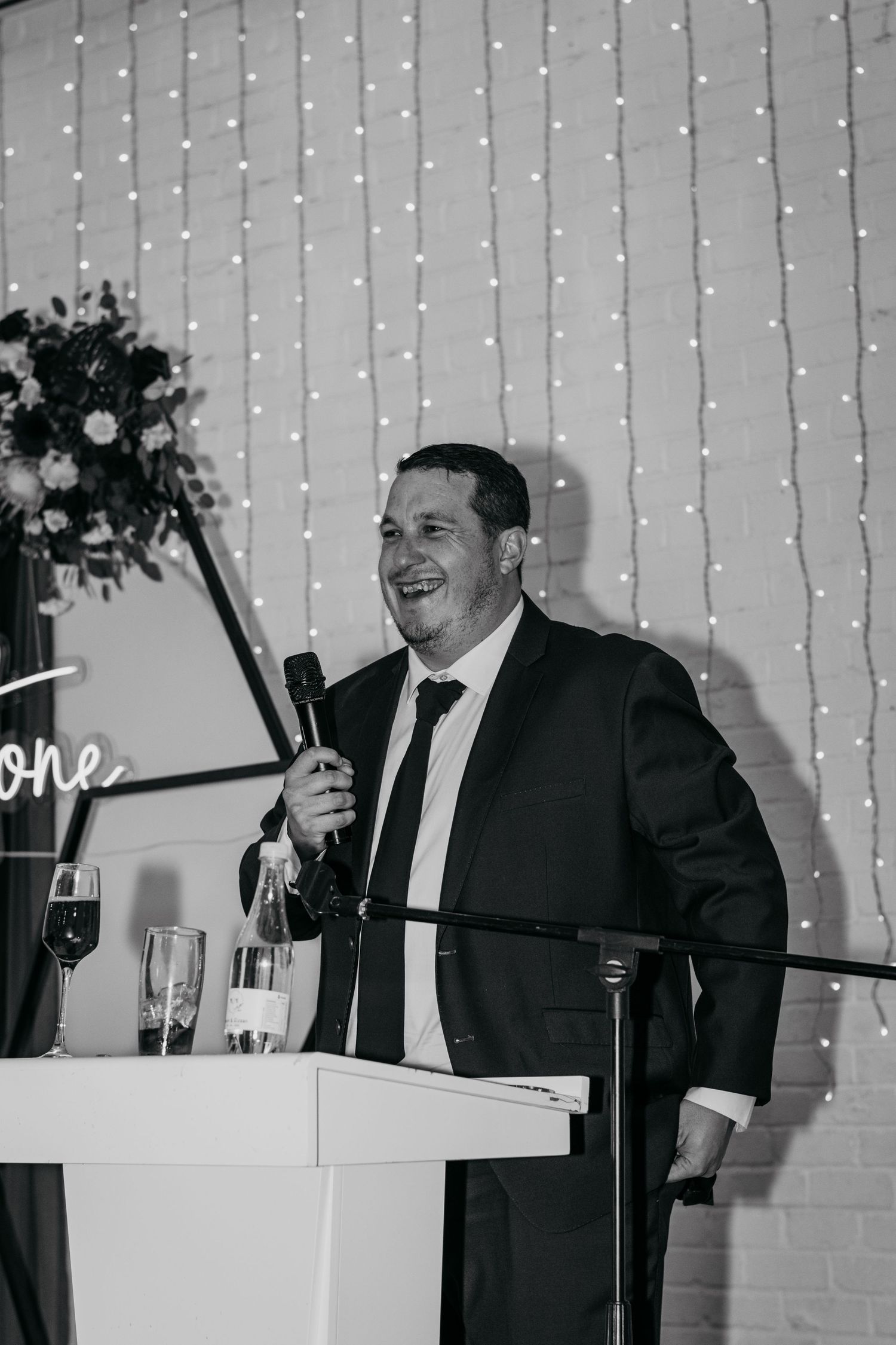 Man in suit giving speech at wedding reception podium with fairy light backdrop in black and white photo.