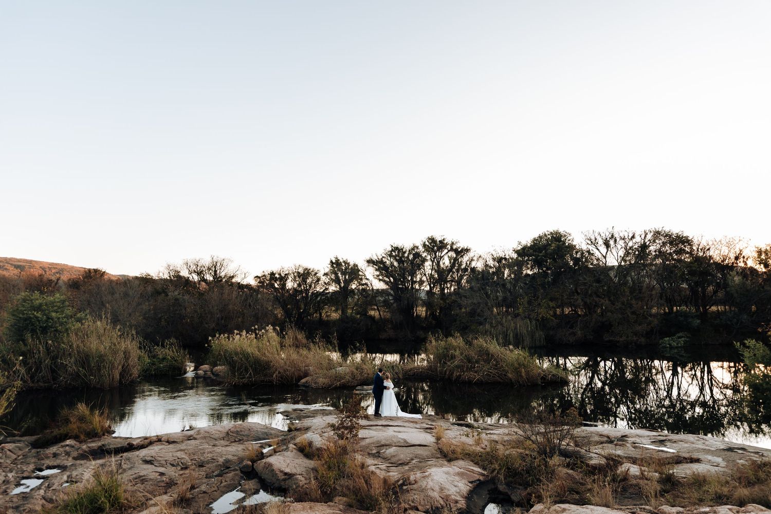 Couple stands on rocky shore beside a tranquil river surrounded by trees at dusk in a scenic outdoor wedding photo.