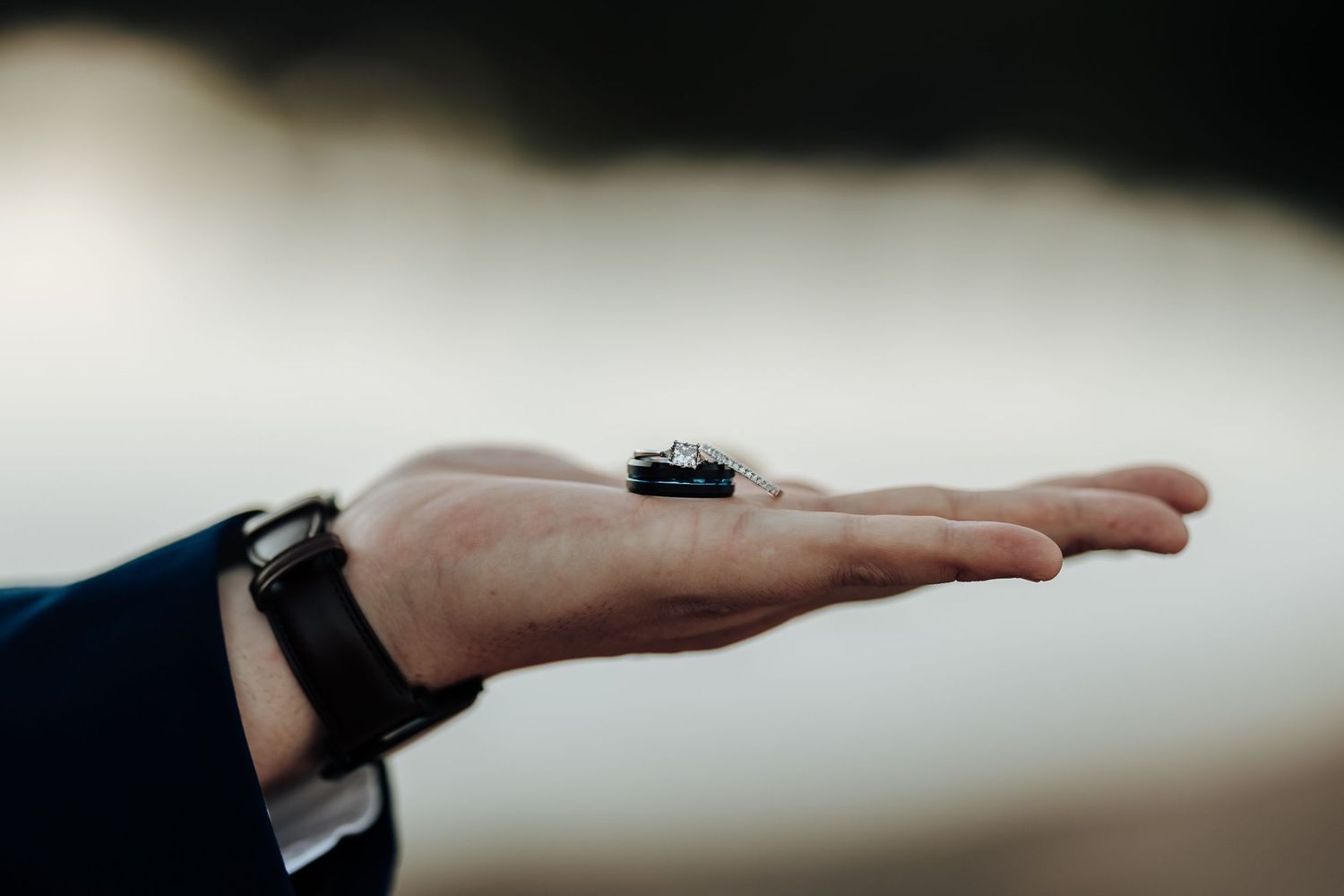 A man's hand wearing a black watch holds a small car key against a blurred outdoor background.