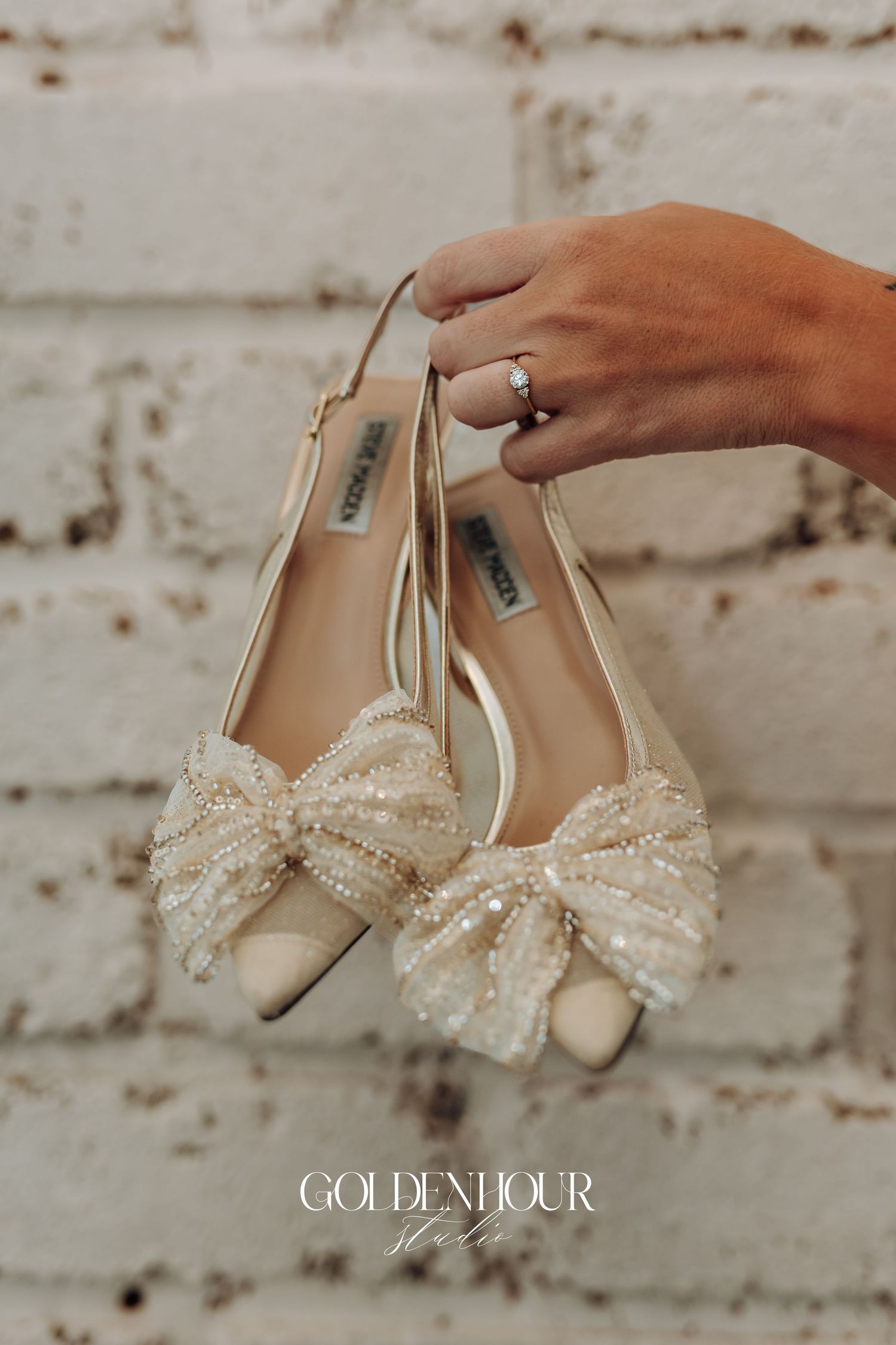 Elegant ivory bridal shoes with pearl and crystal bow details displayed against a rustic white brick wall.
