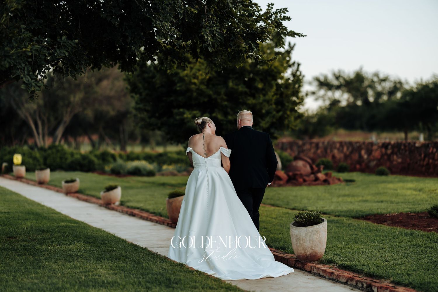 A couple in formal wedding attire embraces while walking along a garden pathway at sunset.