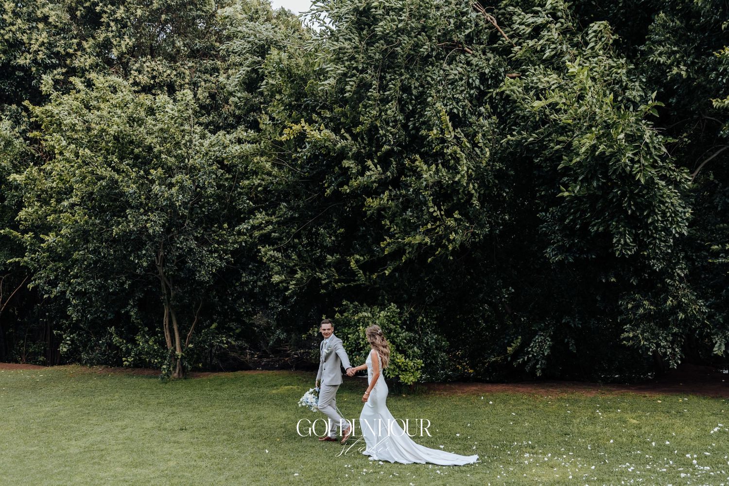 Wedding couple walking together on lush green lawn surrounded by trees.
