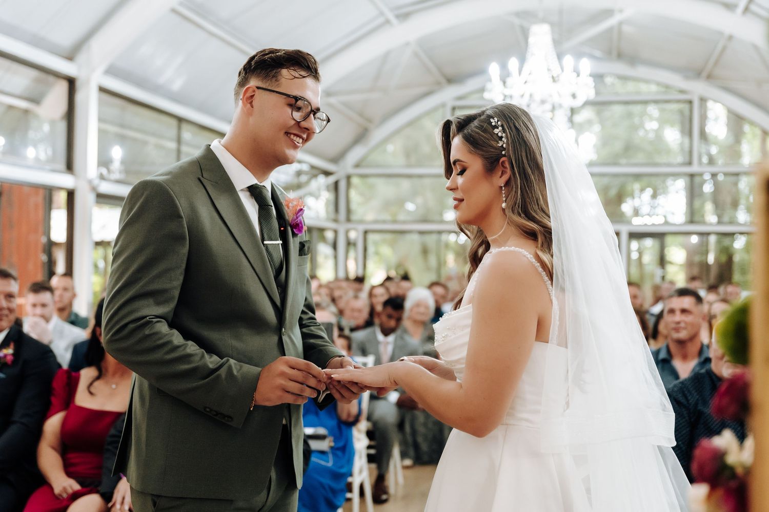 A couple exchanges rings during their wedding ceremony in an elegant greenhouse venue with chandeliers.