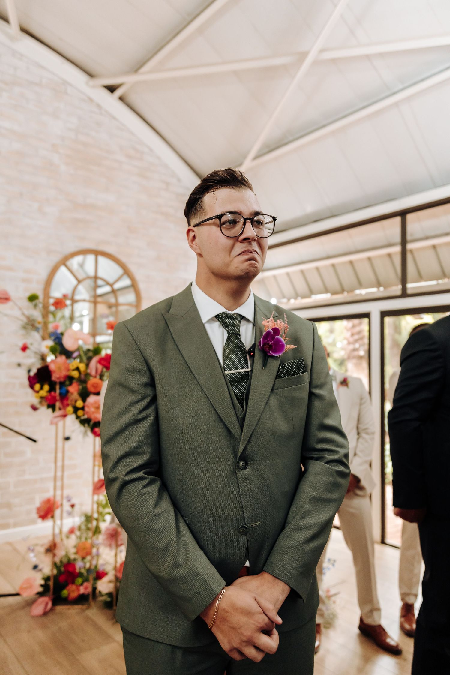 A groom in an olive green suit and glasses stands solemnly during a wedding ceremony with floral decorations.