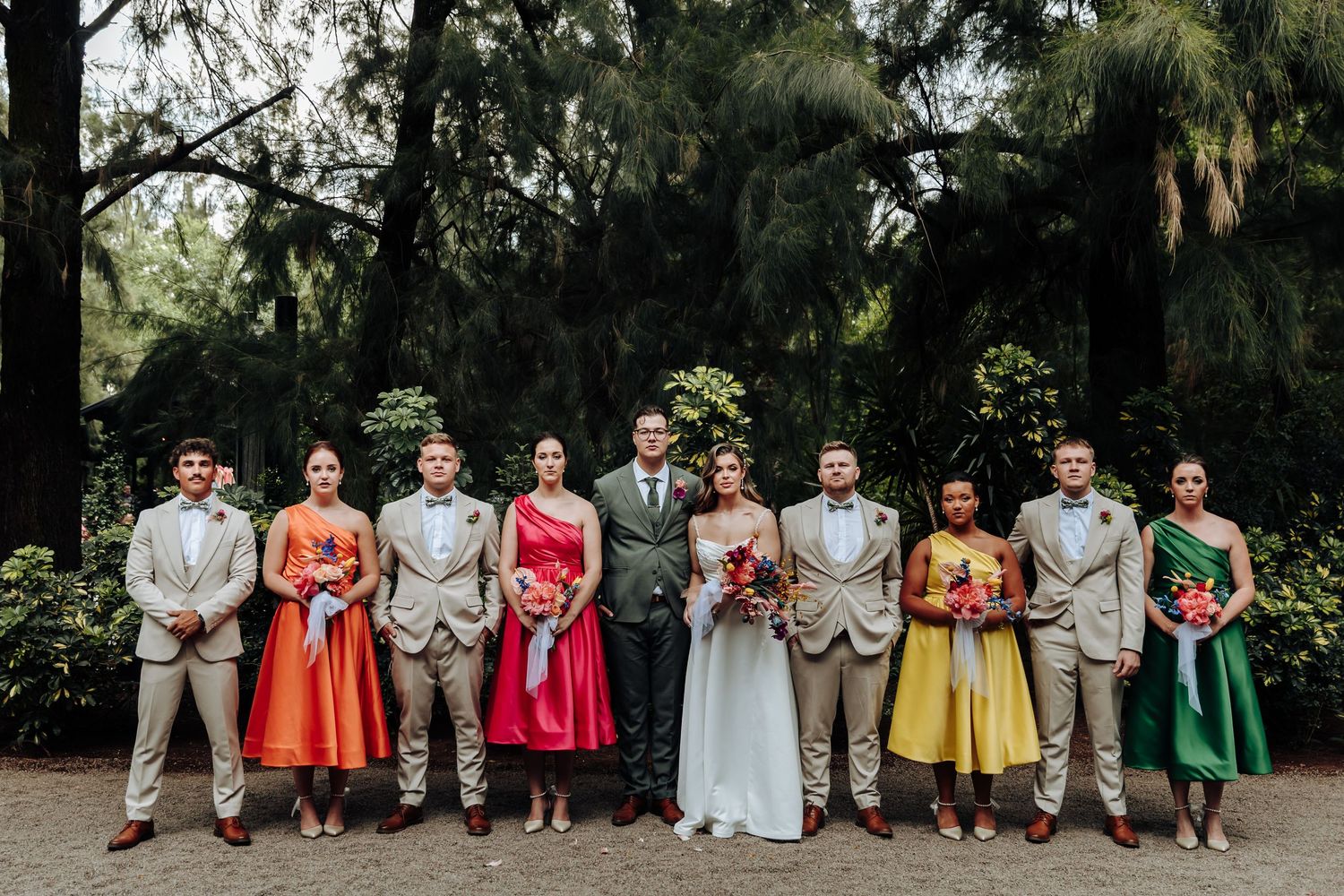 A wedding party stands in a line wearing colorful vintage-style dresses and suits under trees in a garden setting.