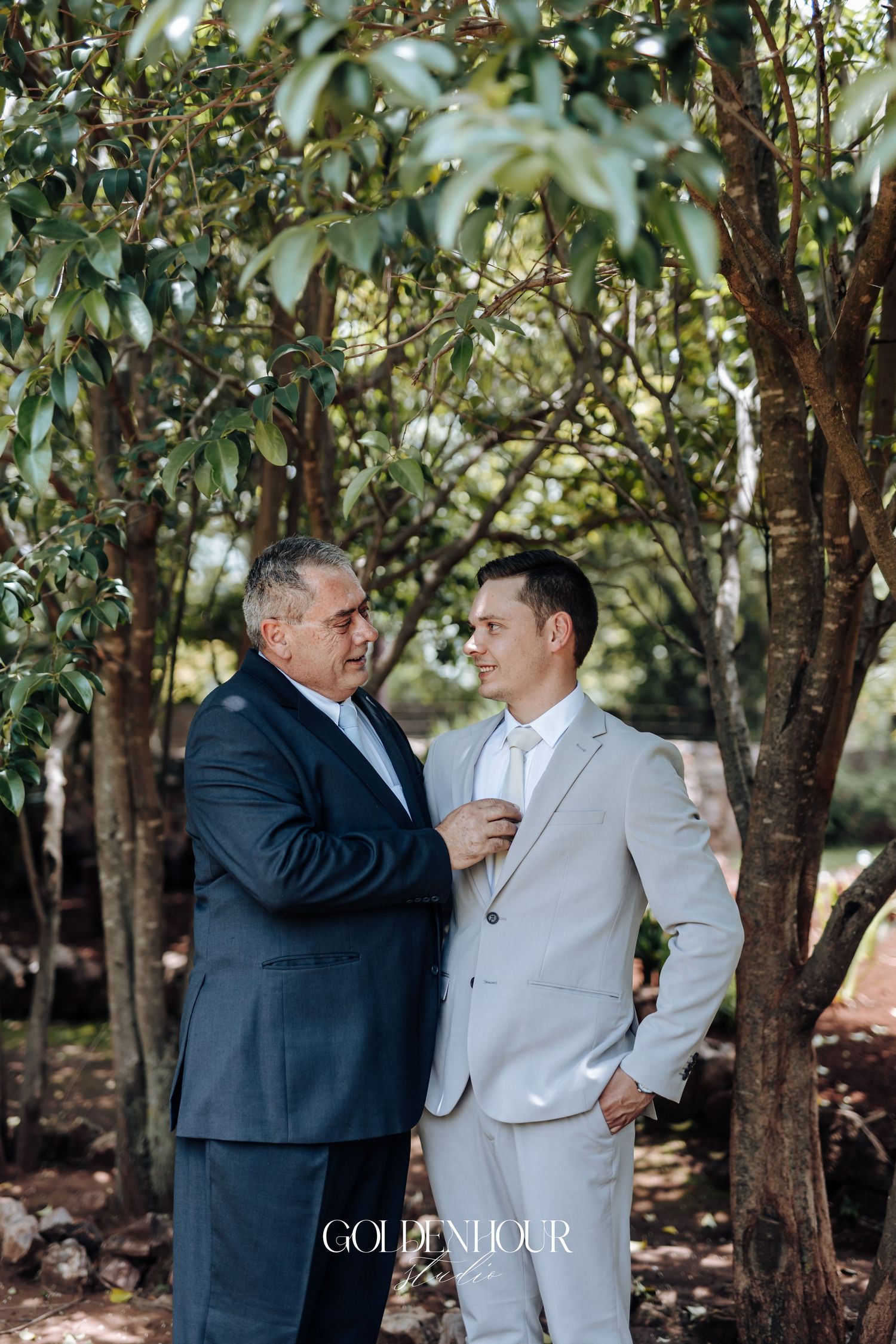 Two men share an emotional embrace in a garden setting with lush trees in the background.