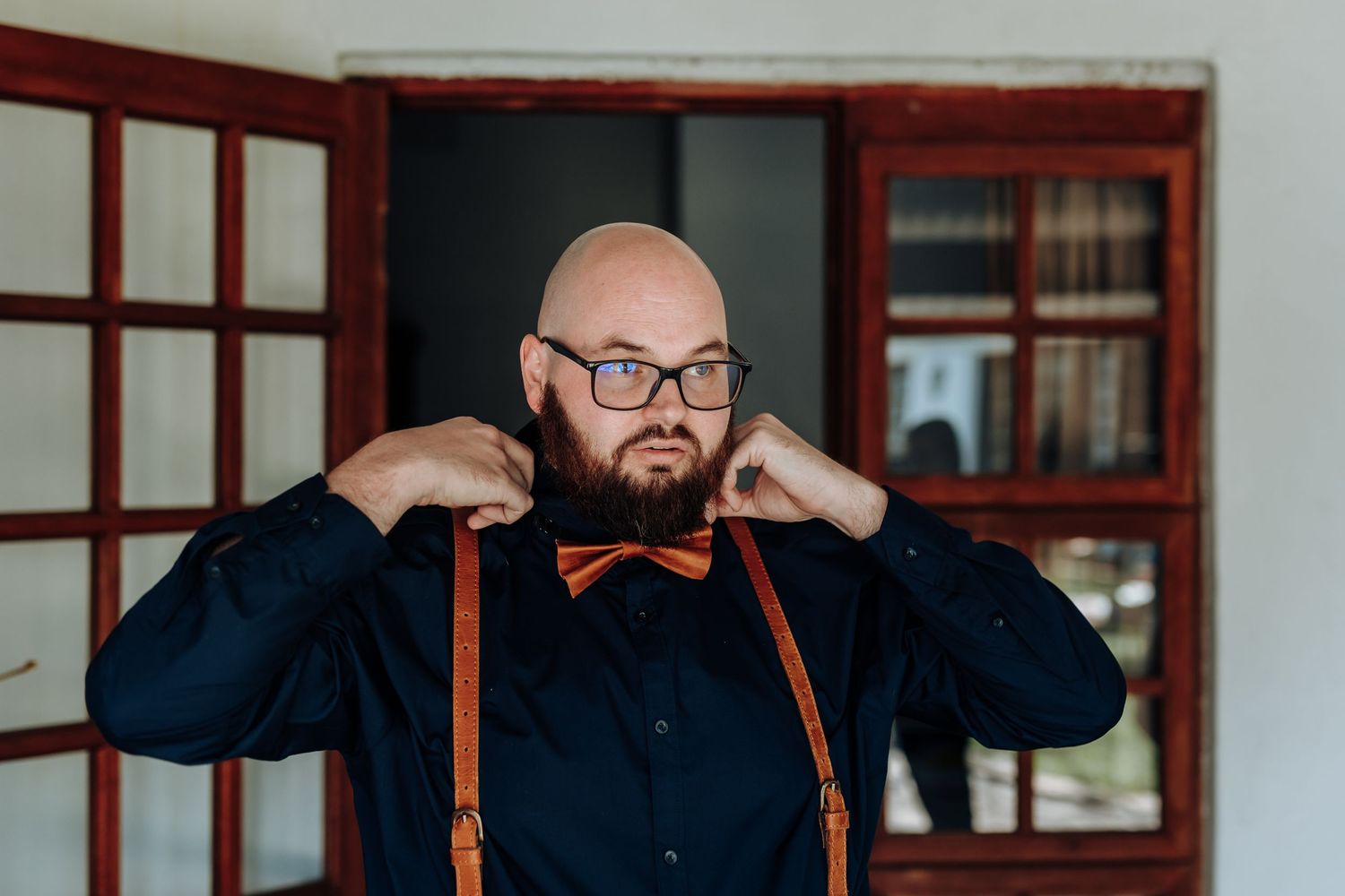 A person wearing glasses adjusts their orange bow tie and brown leather suspenders while wearing a navy blue shirt.