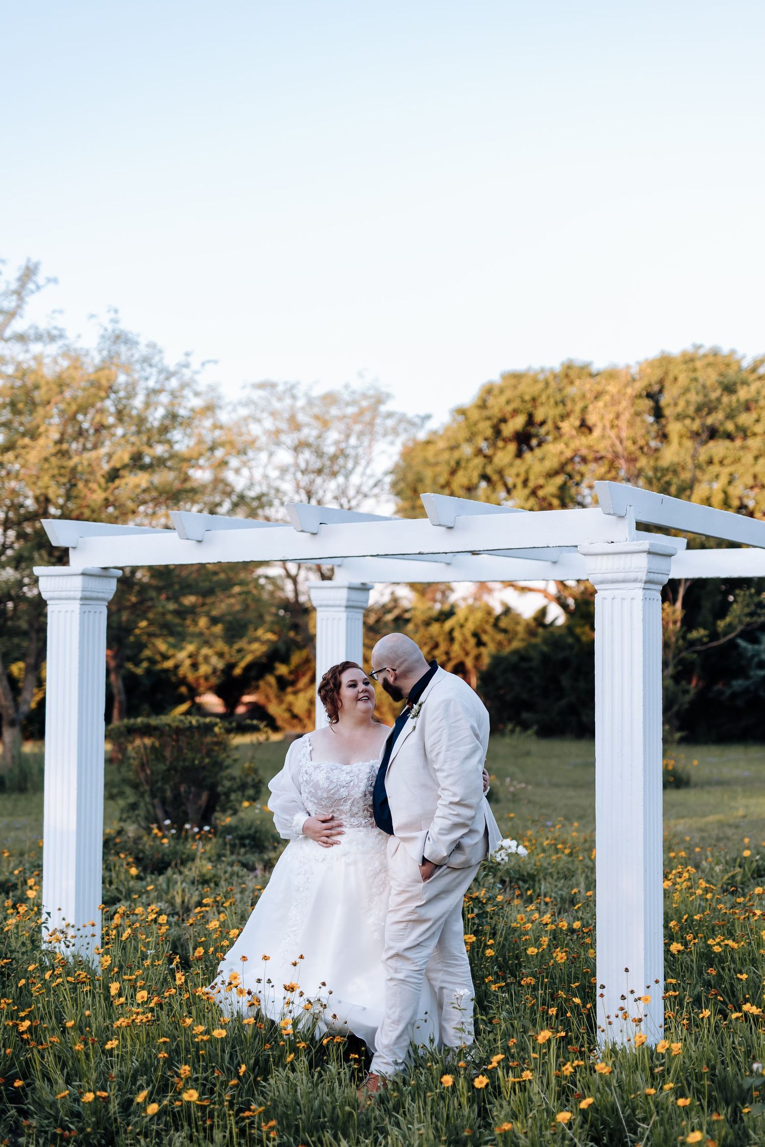 A romantic couple shares tender moments in a wildflower meadow with white pergola columns at sunset.
