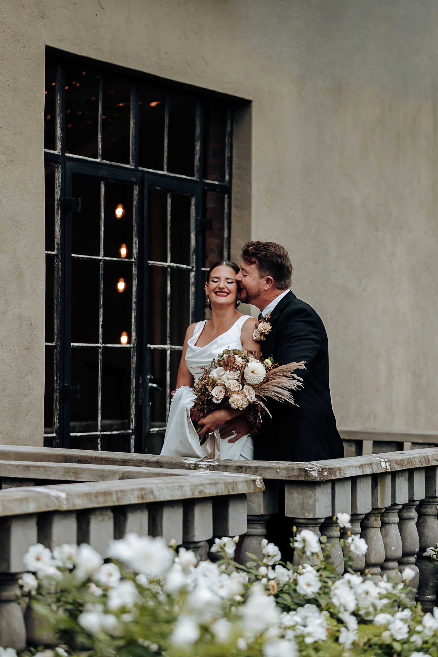 Romantic wedding couple embraces on an elegant stone balcony surrounded by delicate white flowers.
