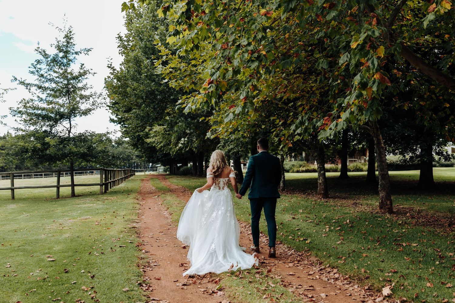 A couple walks down a tree-lined dirt path in a park setting, with the bride wearing a flowing white gown.