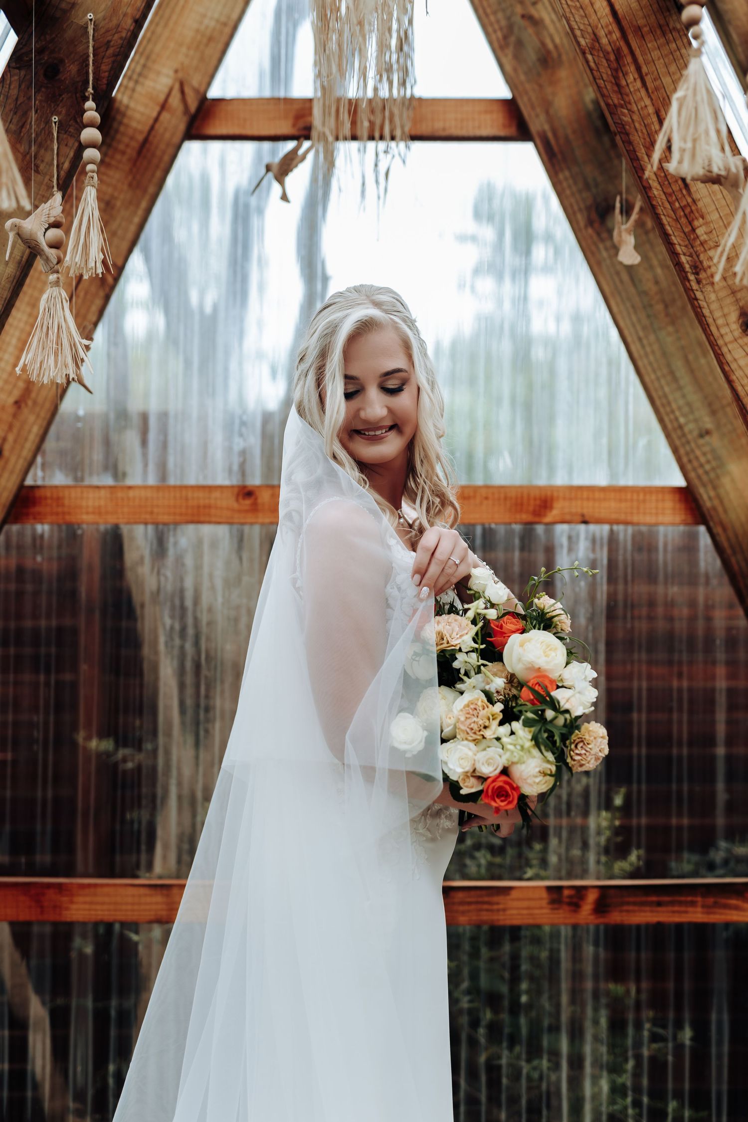 Elegant bride in flowing white dress holds a romantic bouquet of roses and mixed flowers in a rustic wooden gazebo.