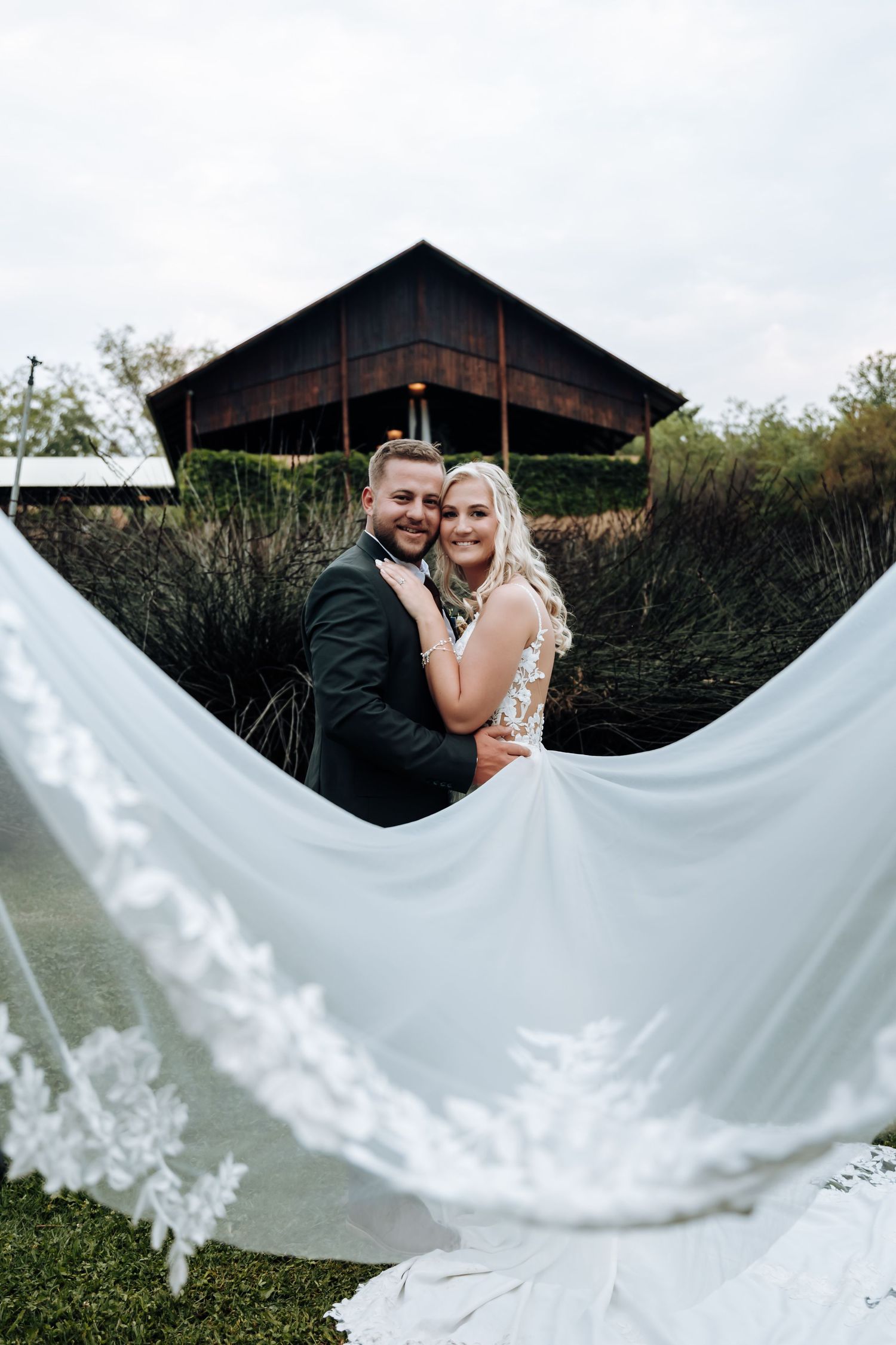 A wedding couple poses in front of a rustic barn while the bride's lace-trimmed dress train flows in the foreground.
