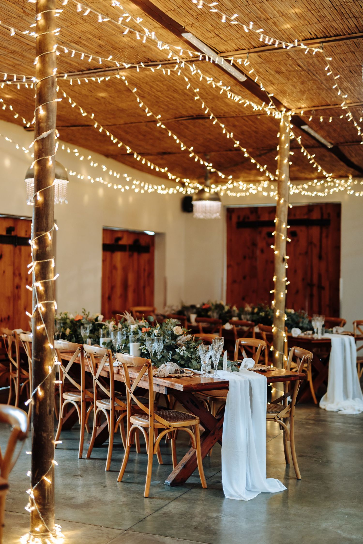 Rustic wedding reception venue decorated with string lights and wooden tables arranged in rows beneath exposed beams.