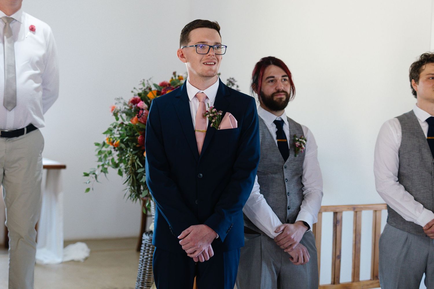 People gather for a wedding ceremony in a bright indoor venue with decorative floral arrangements.