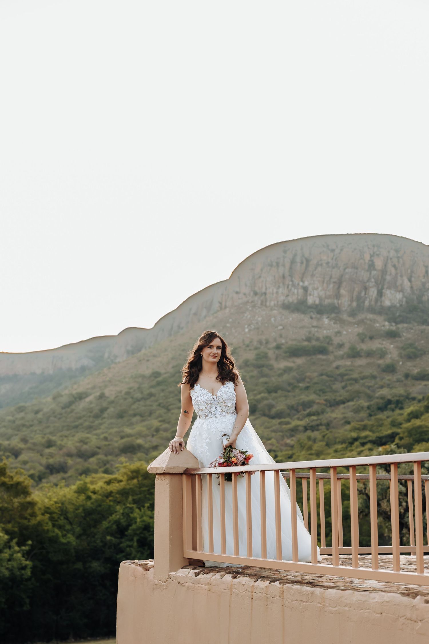 A bride in a white dress poses on a scenic balcony overlooking mountains and greenery in the background.