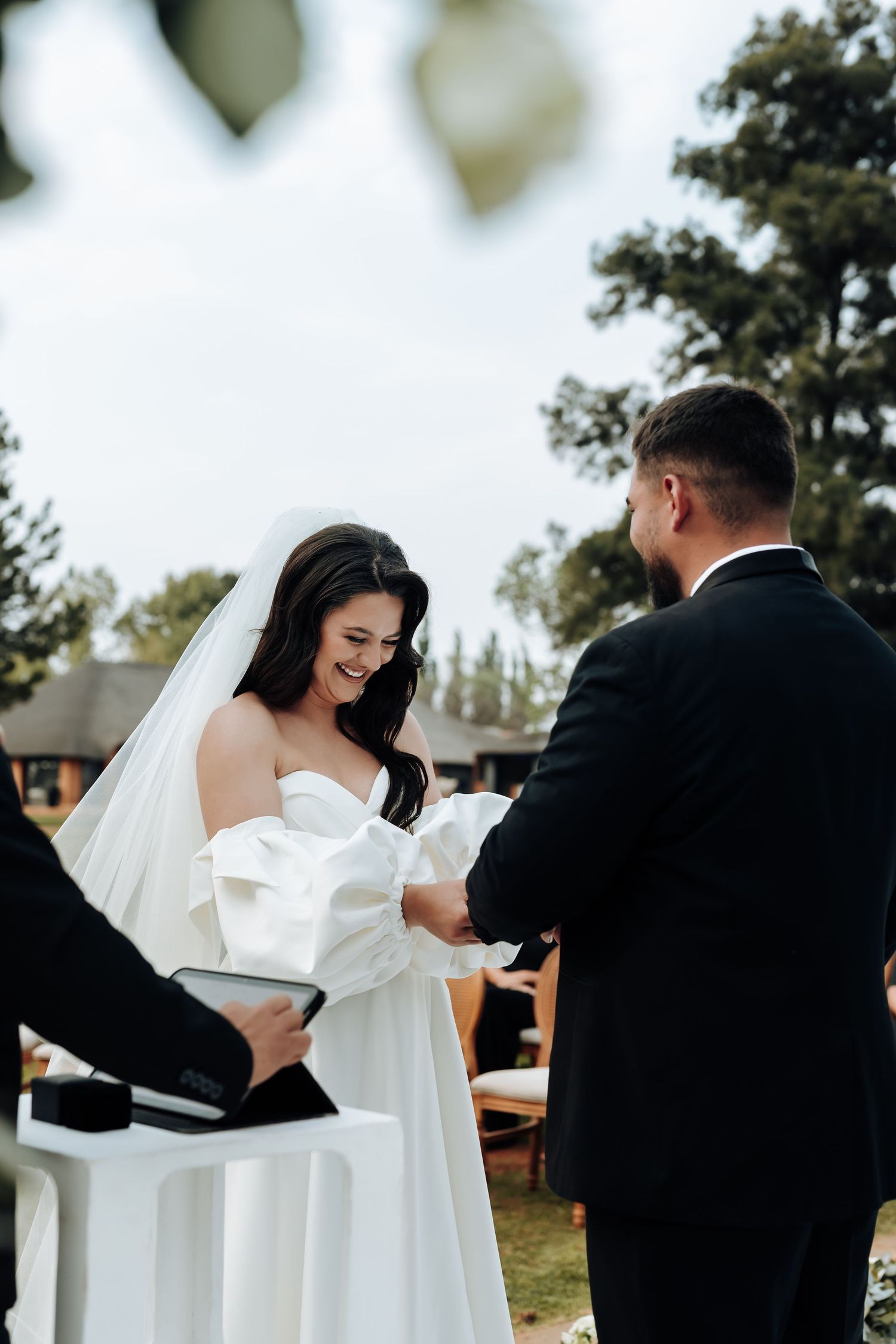 A bride in a white wedding dress embraces someone in a black suit during an outdoor ceremony.