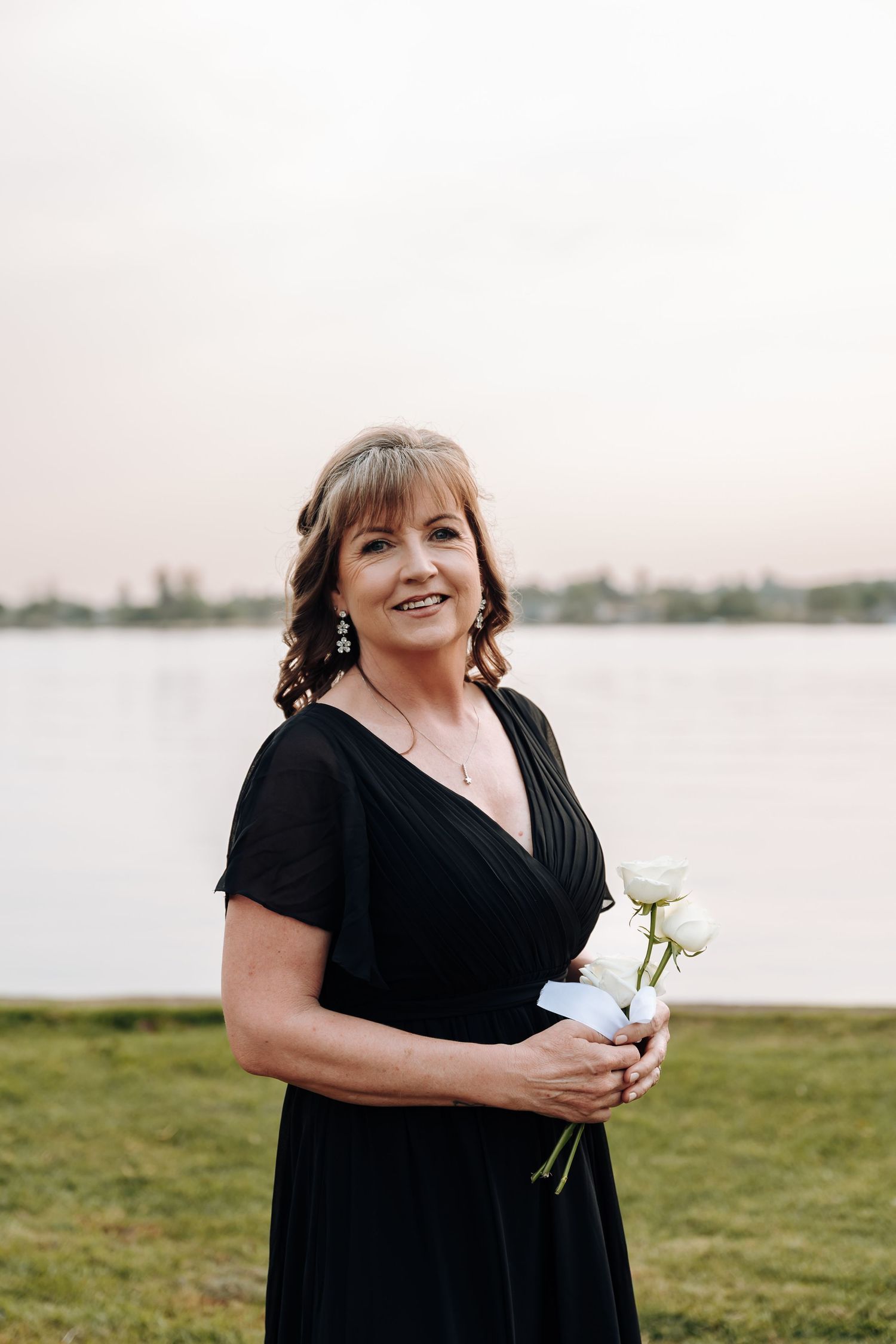 Person in black dress holding flowers stands by waterfront at sunset with joyful expression.