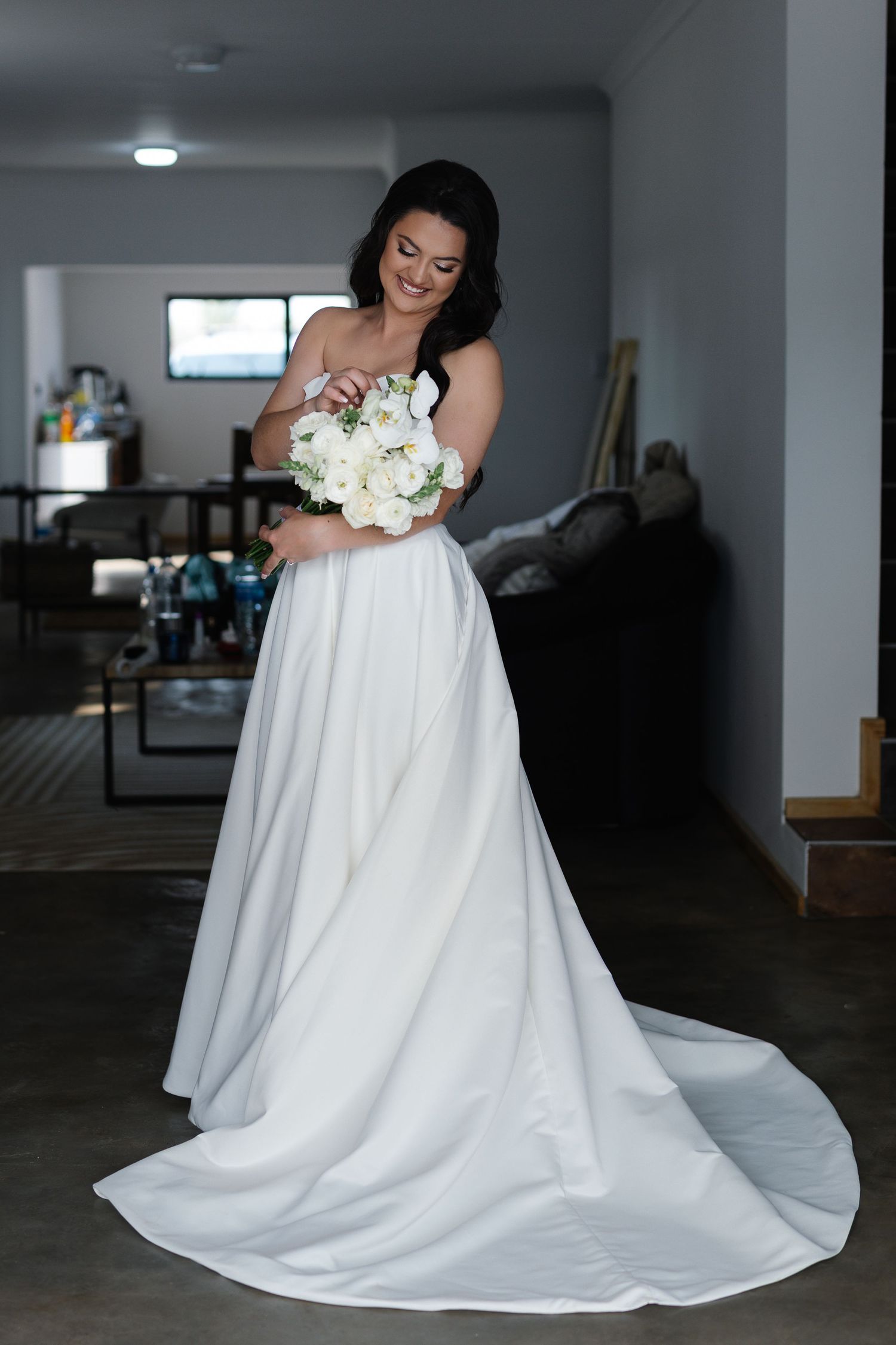A bride in a flowing white wedding gown holds a bouquet of white flowers in a dimly lit indoor setting.