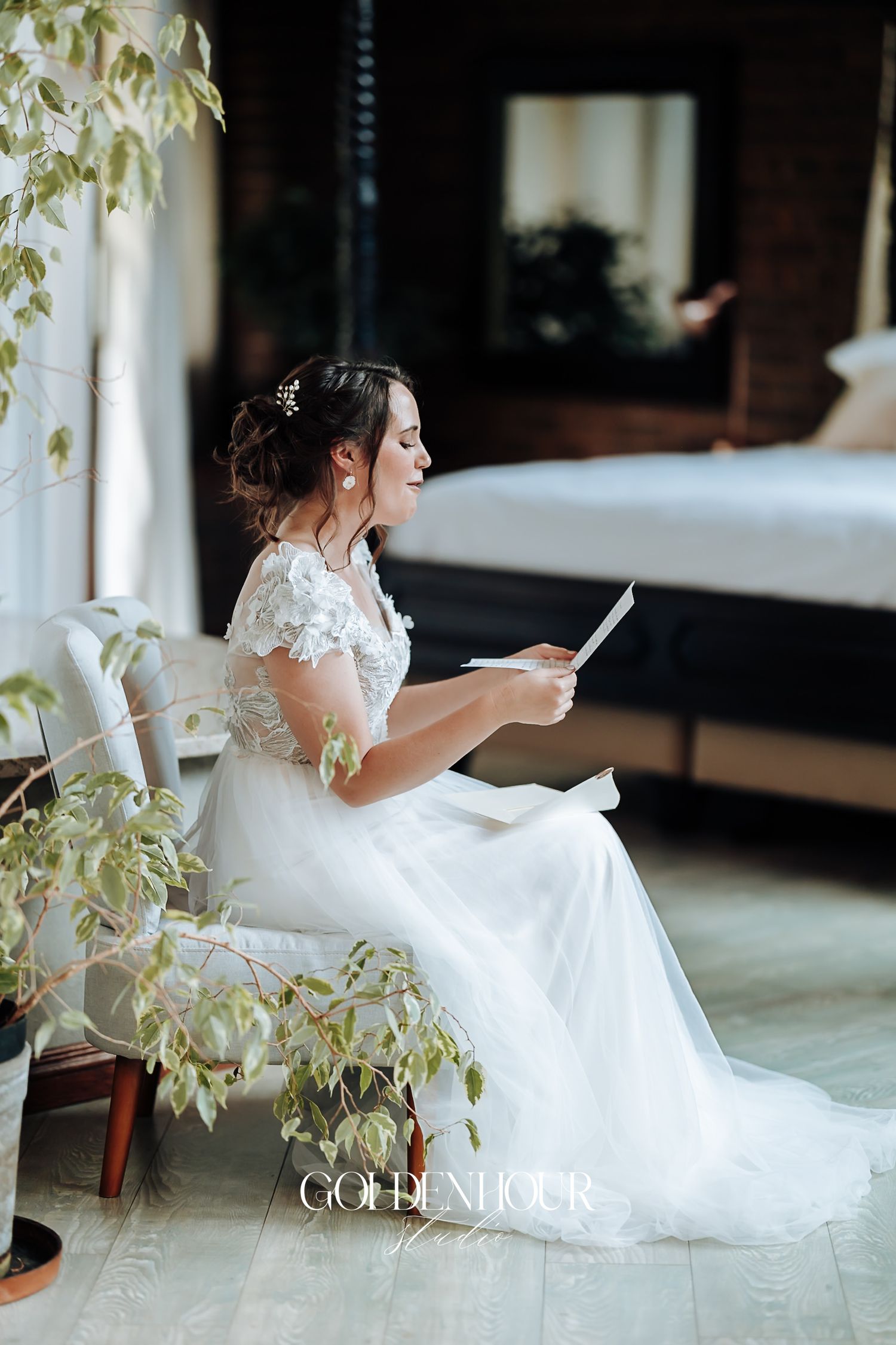 Bride in lace wedding gown reads a letter while seated by lush greenery in an elegant room.