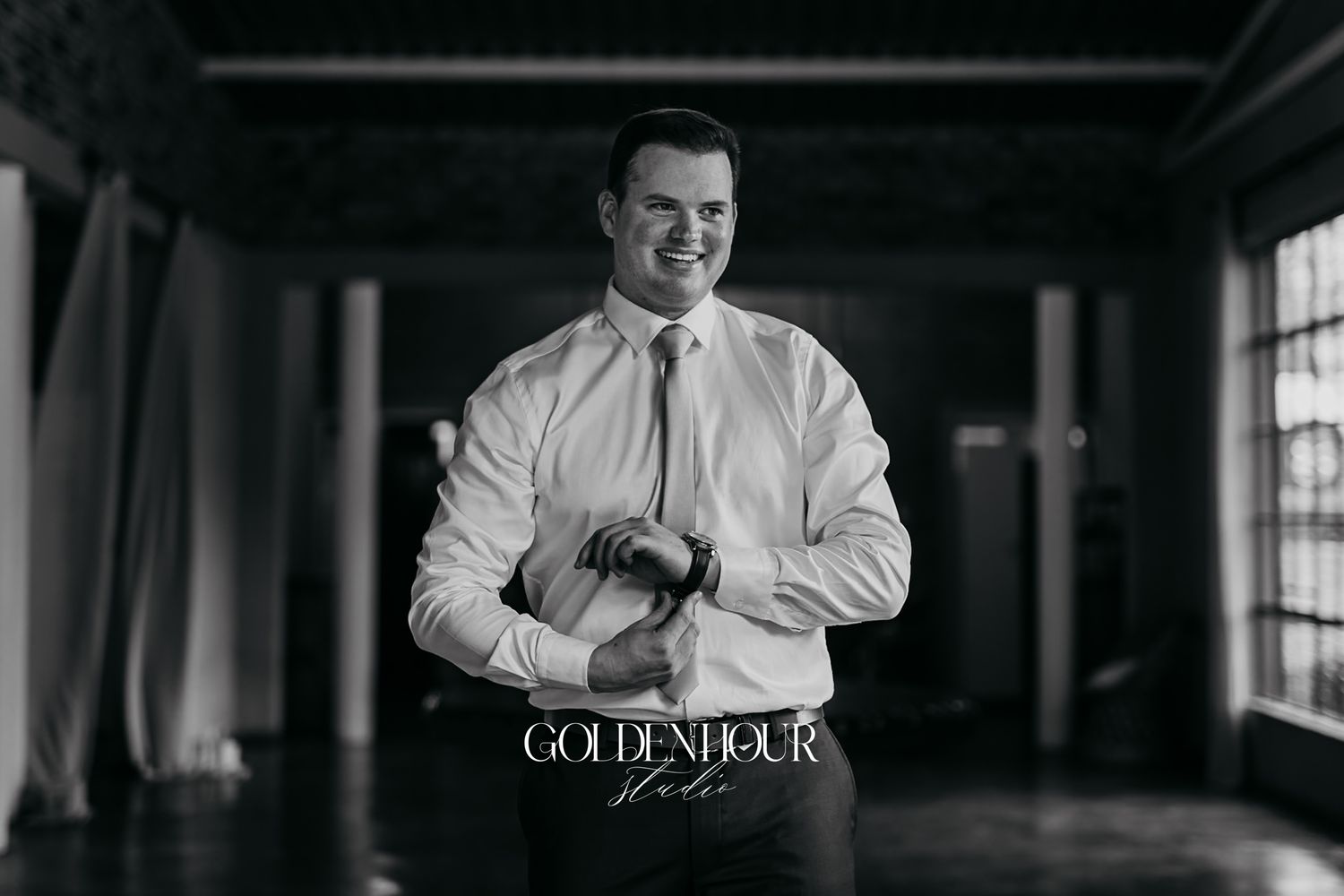 Black and white photo of a groom adjusting his cufflinks in a bright hallway, smiling confidently on his wedding day.