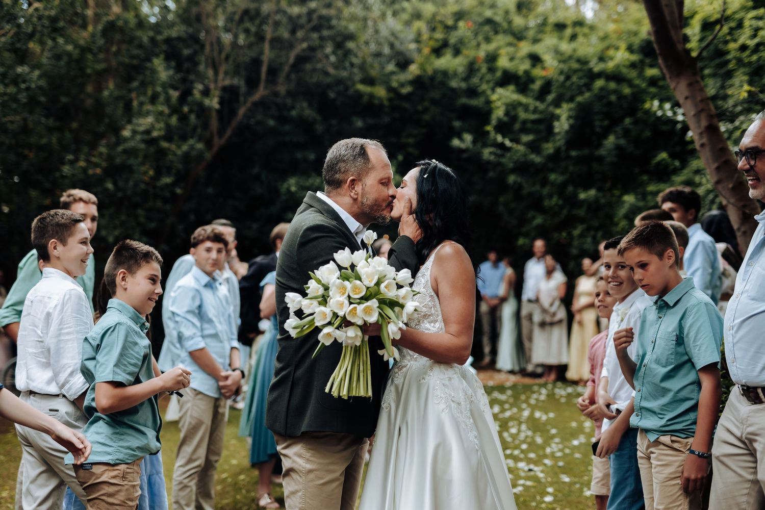 Bride and groom share first kiss at outdoor wedding ceremony surrounded by children in matching teal outfits.
