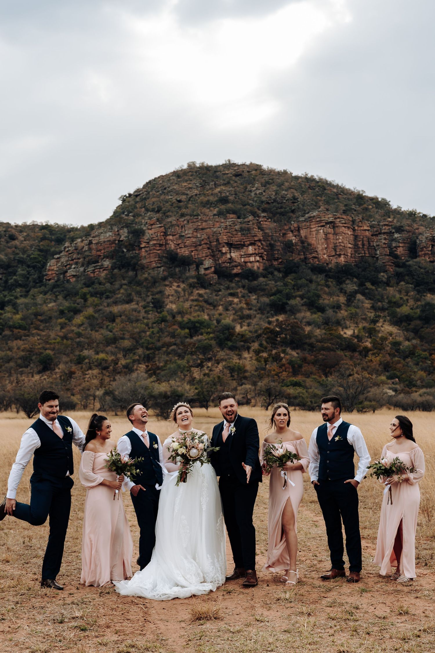 Wedding party poses in front of dramatic rocky mountain backdrop in arid landscape during outdoor celebration.