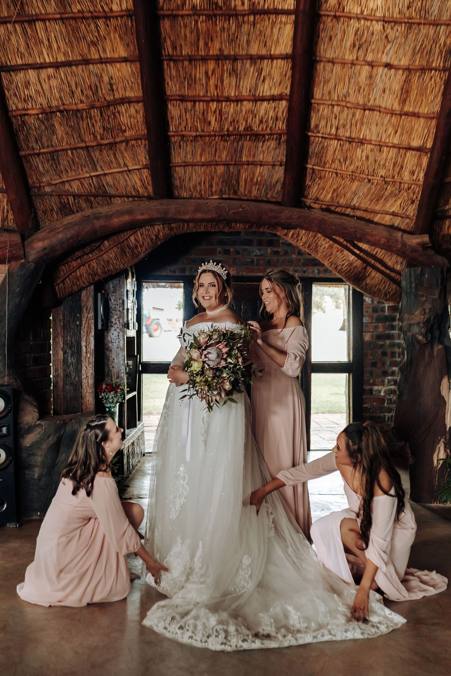 Bride in elegant white lace gown gets help from bridesmaids in blush dresses under rustic thatched roof venue.