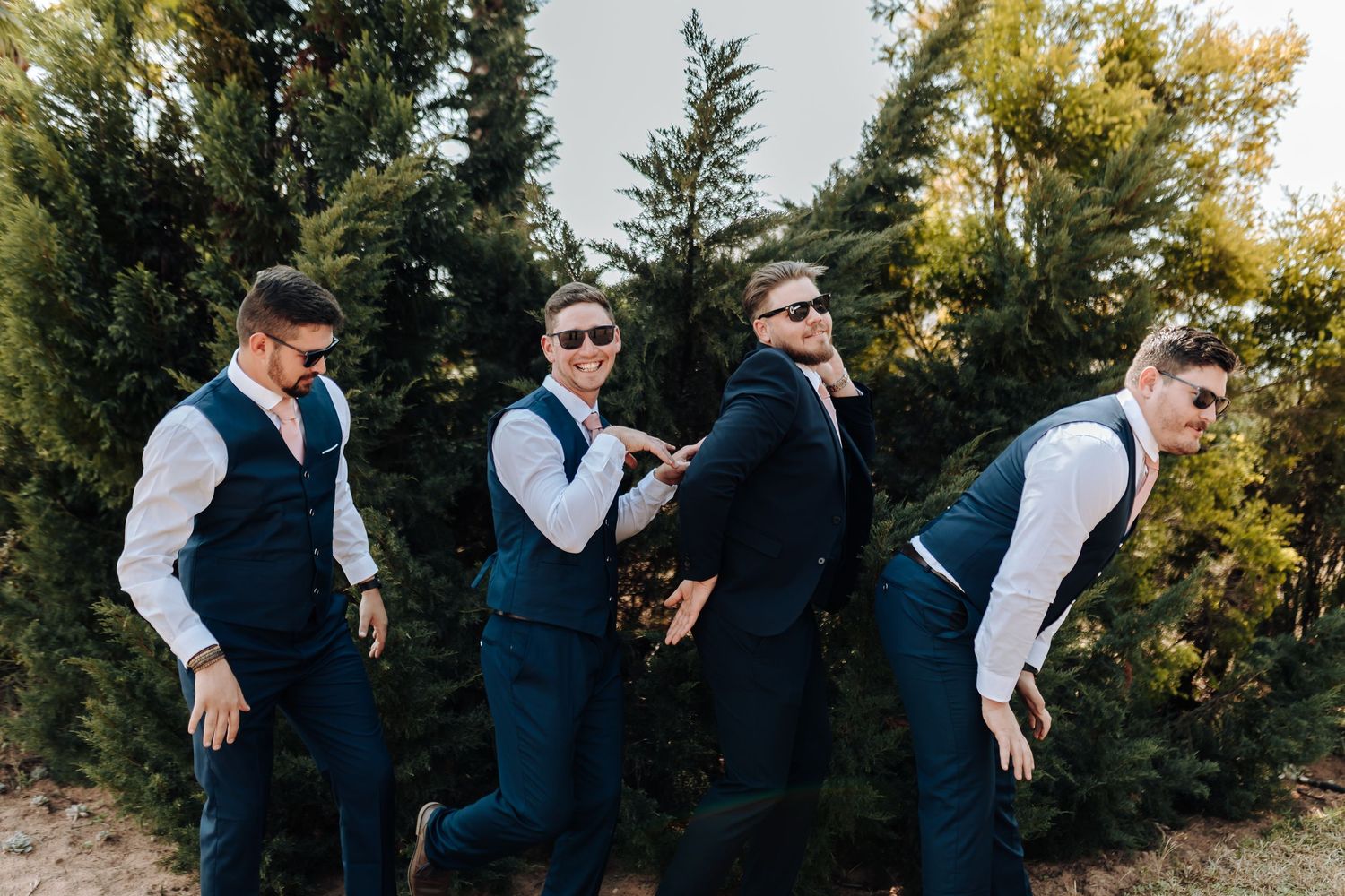 Four groomsmen in matching navy vests and white shirts strike playful poses in an outdoor setting with trees.