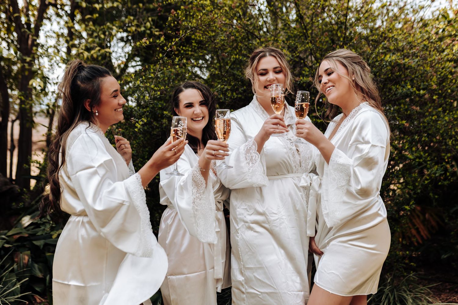 Four people in white robes laughing and drinking beverages together during a celebratory gathering outdoors.