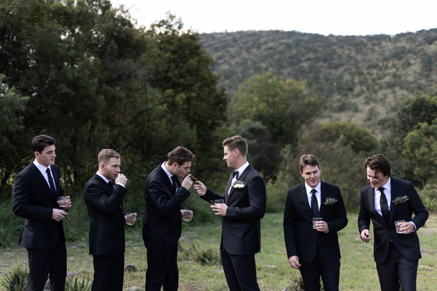 Group of groomsmen in dark suits smoking cigars against a hillside backdrop.