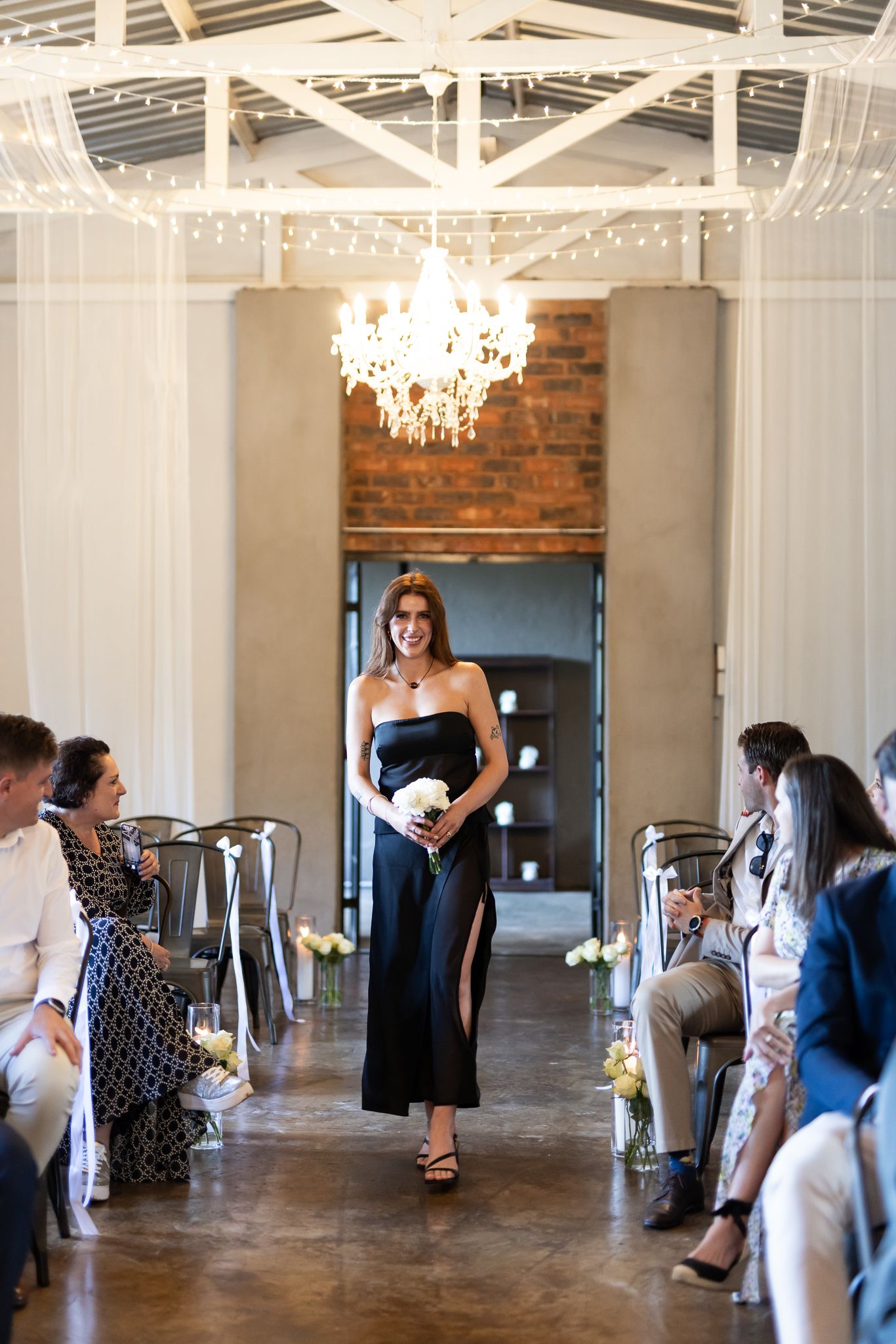 Bridesmaid walks down aisle in black dress holding white bouquet under elegant chandelier.