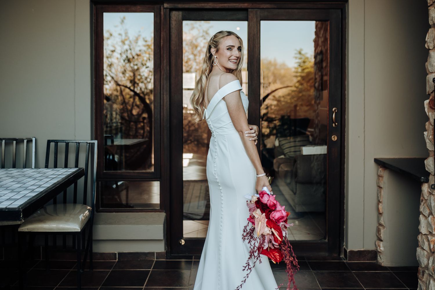 Elegant bride in white off-shoulder gown poses with vibrant red bouquet against glass doors at sunset.