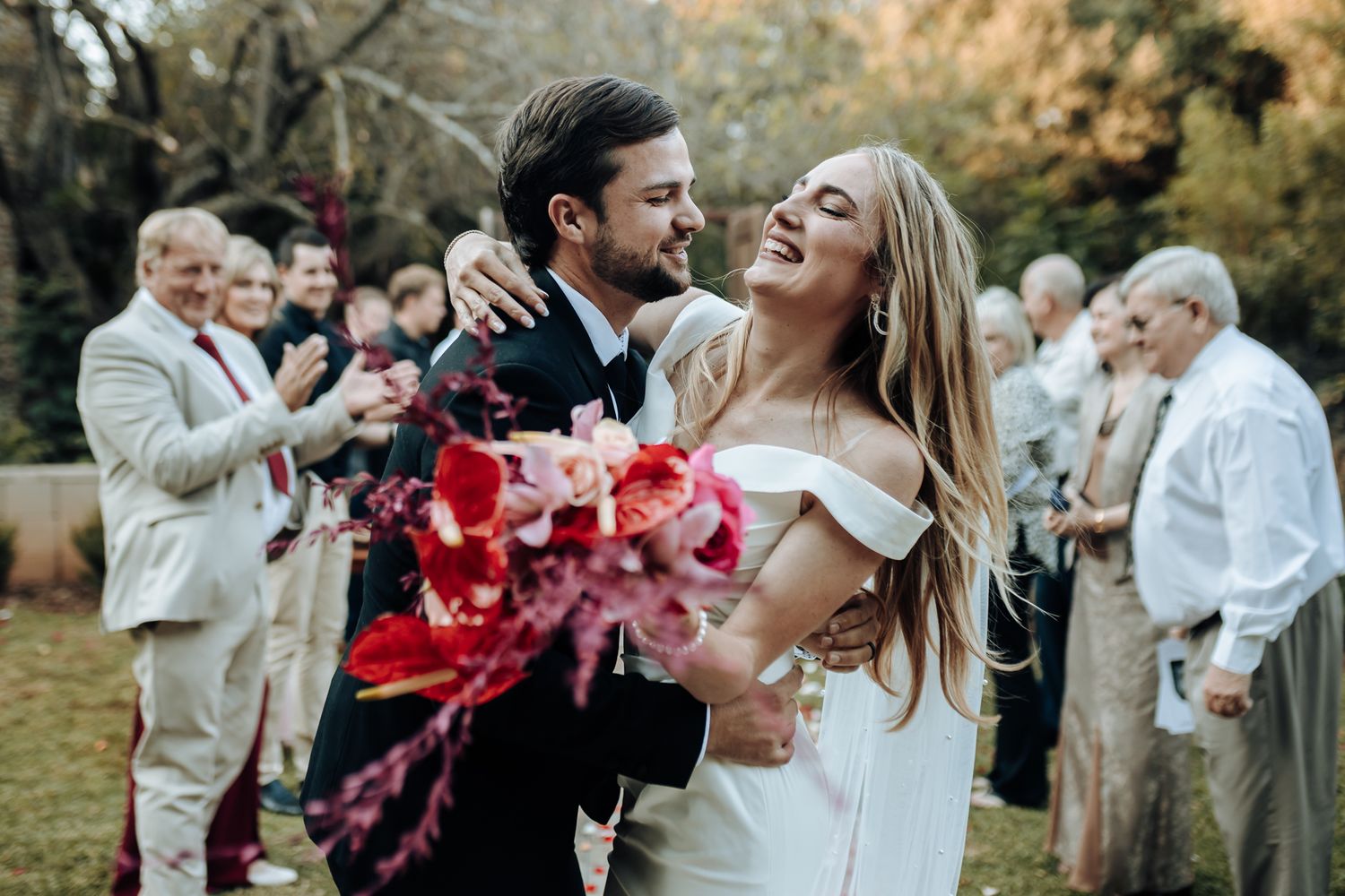 Newlyweds share celebratory moment with wedding guests cheering in background during outdoor ceremony.
