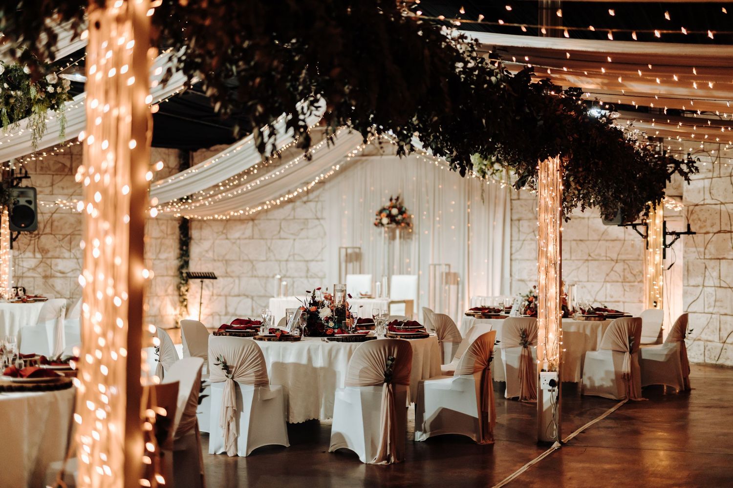 Wedding reception venue decorated with string lights, white draping, and round tables with elegant place settings.