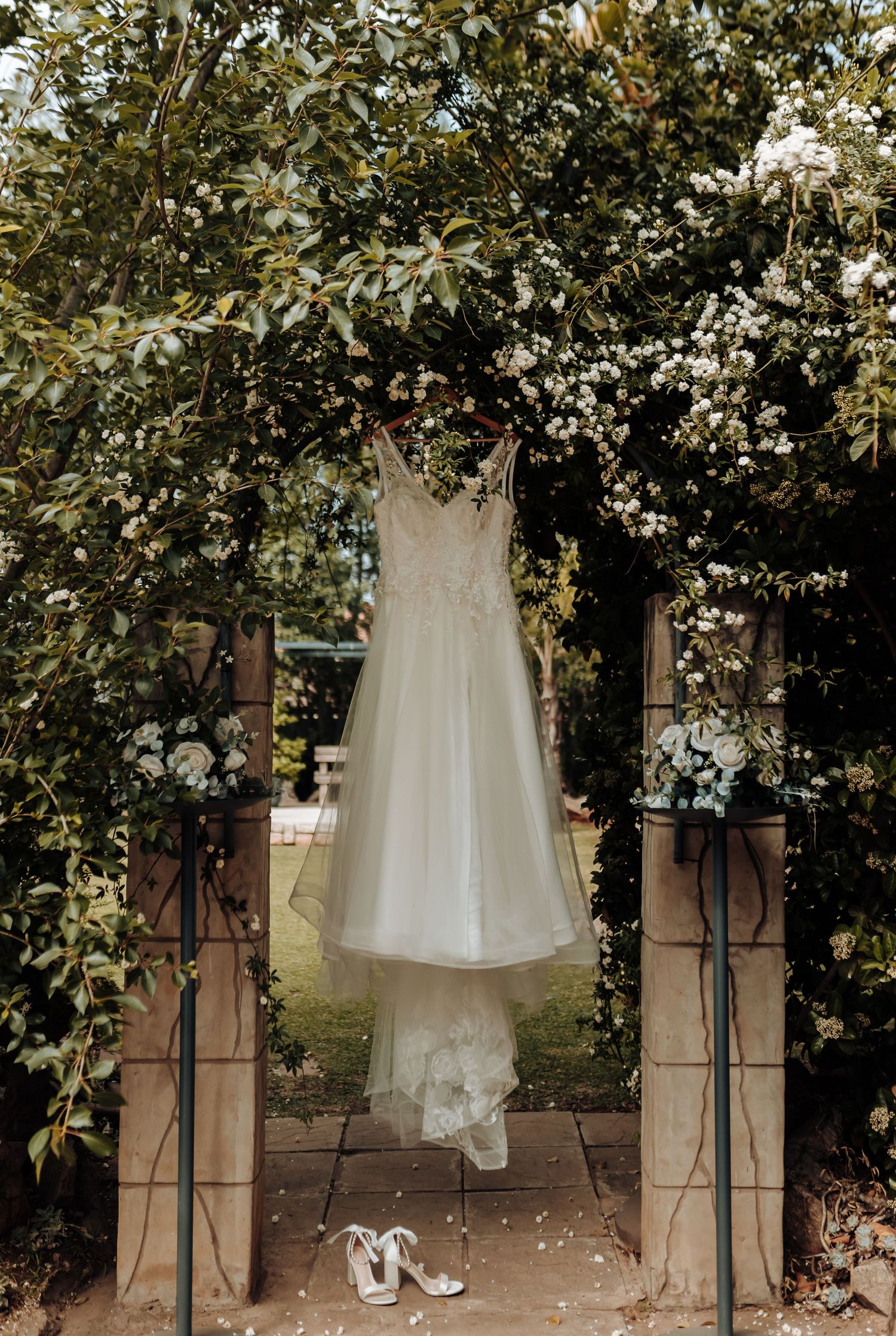 White wedding dress hangs elegantly on garden archway with shoes below.