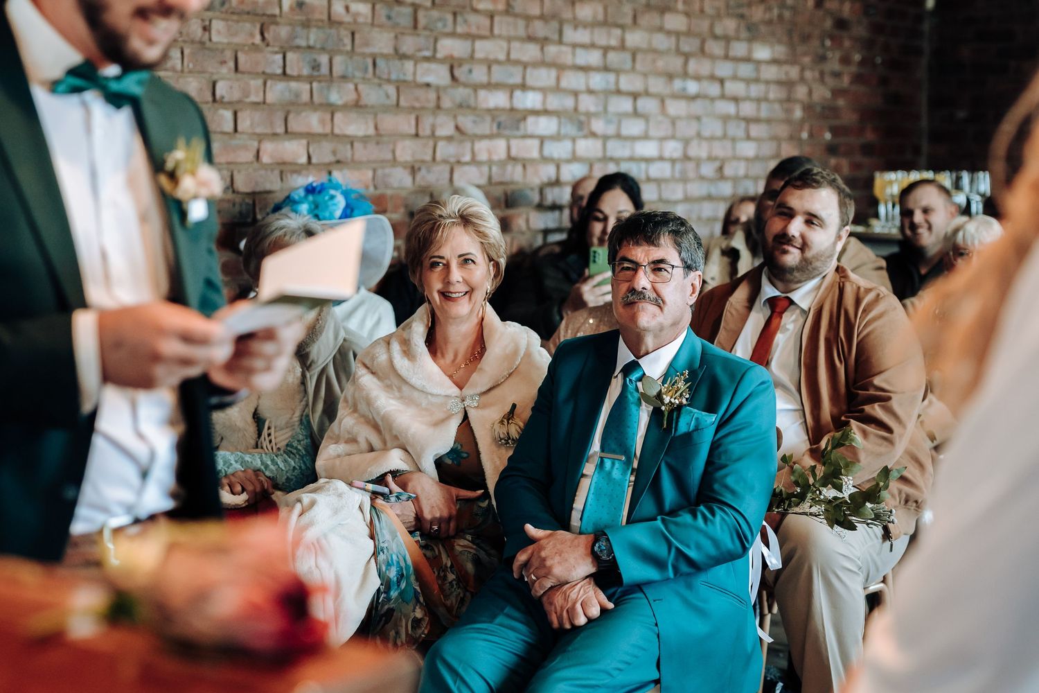 Wedding guests smile and laugh during speeches at a rustic indoor reception with exposed brick walls.