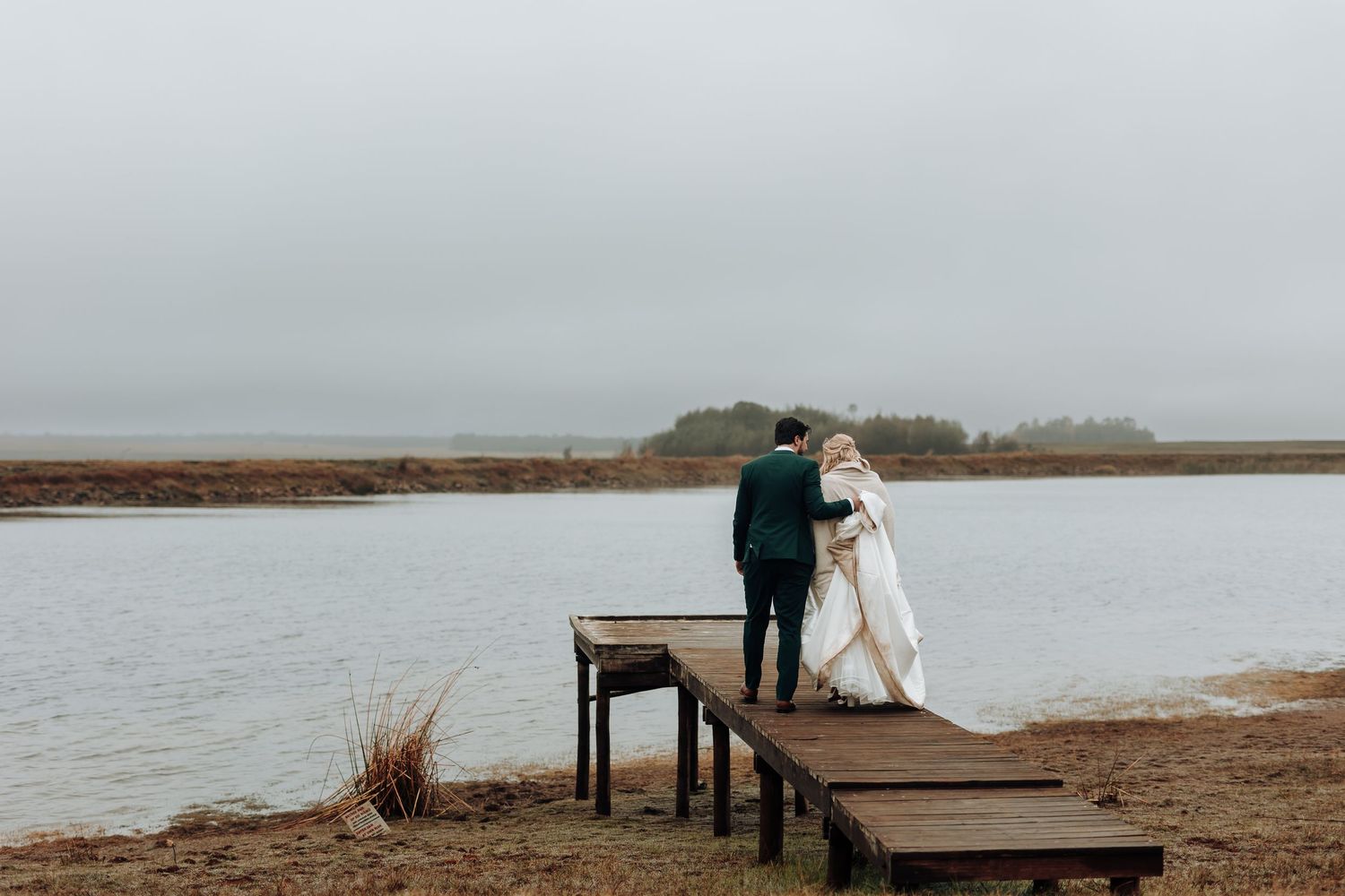 Newlyweds stand together on wooden dock surrounded by coastal scenery.