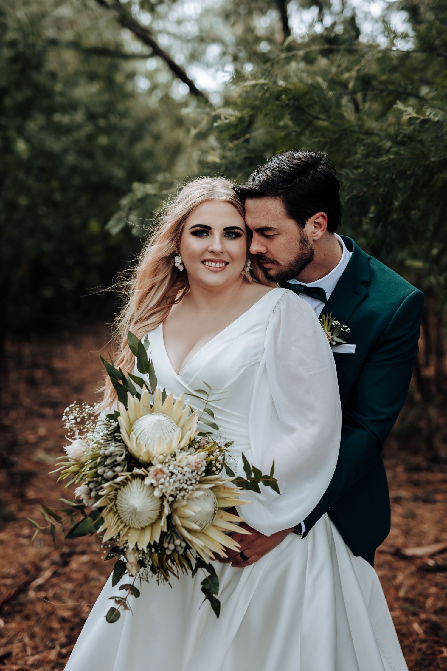 Intimate wedding portrait with emerald suited groom embracing bride holding cream floral bouquet.