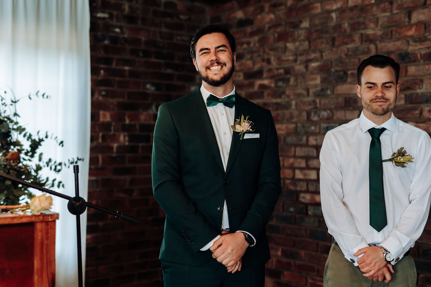 Two groomsmen in formal attire pose against brick wall at wedding ceremony.