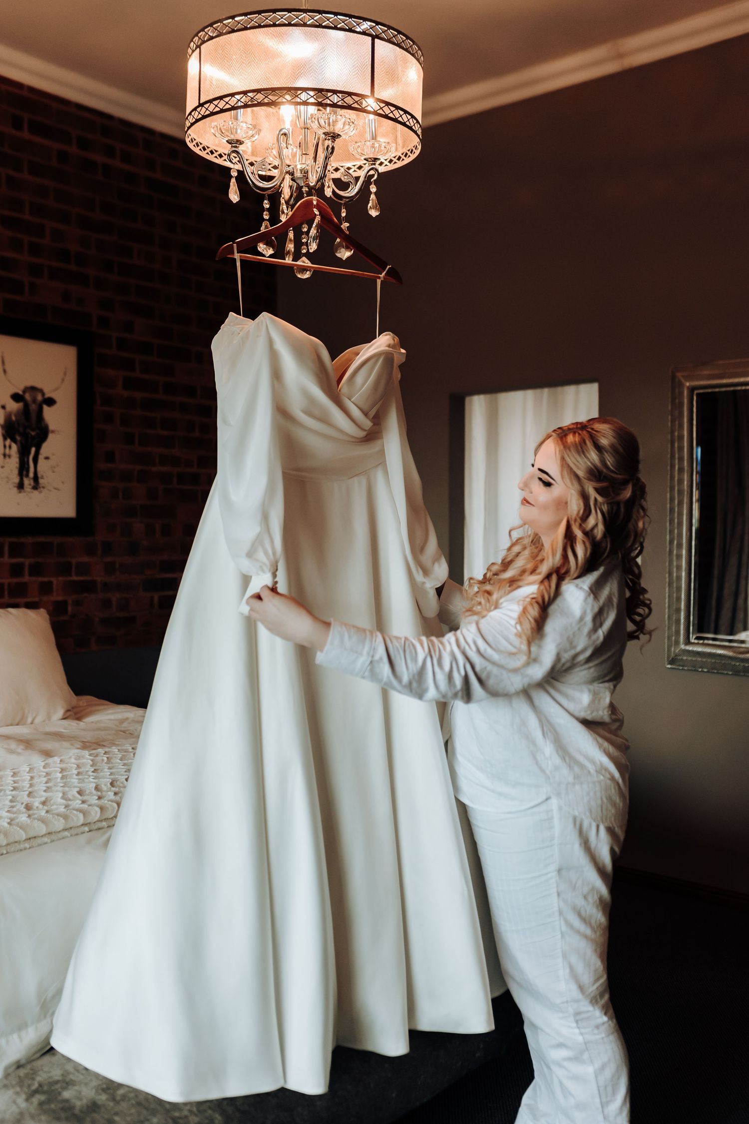 A wedding dress hangs from an elegant crystal chandelier in a room with exposed brick walls.