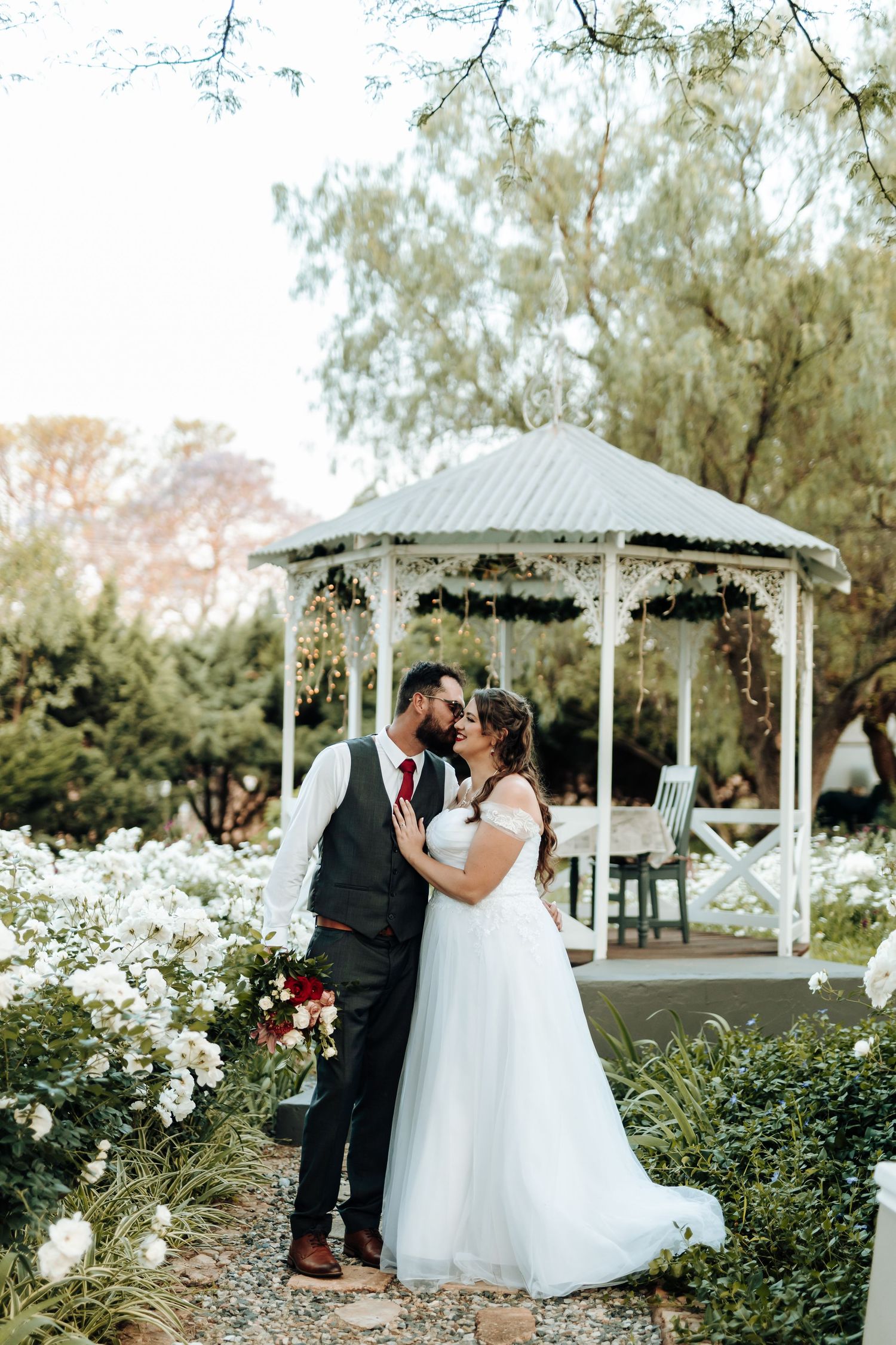 Romantic wedding scene with a couple standing by a decorated white gazebo surrounded by blooming flowers.