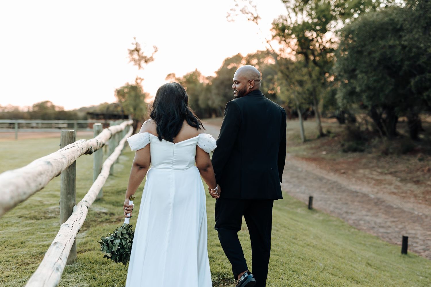 A couple in wedding attire walks hand in hand along a countryside fence at sunset.