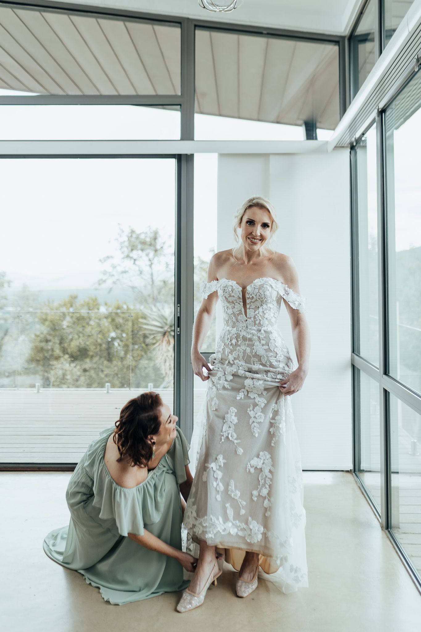 Wedding dress preparations with mint green bridesmaid helping adjust the hem of an intricate lace gown.