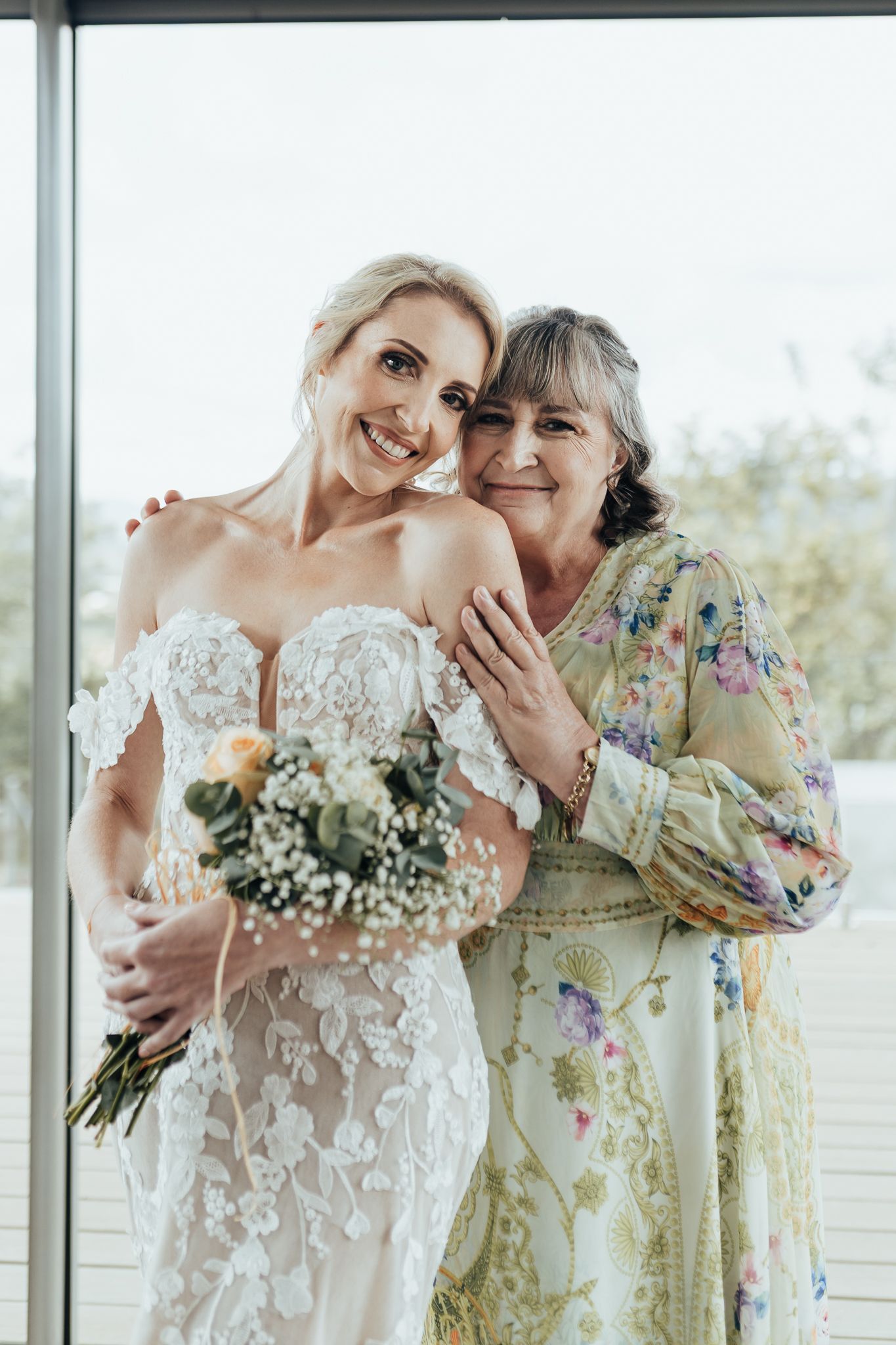 Two figures embracing in formal attire stand by window with floral bouquet.