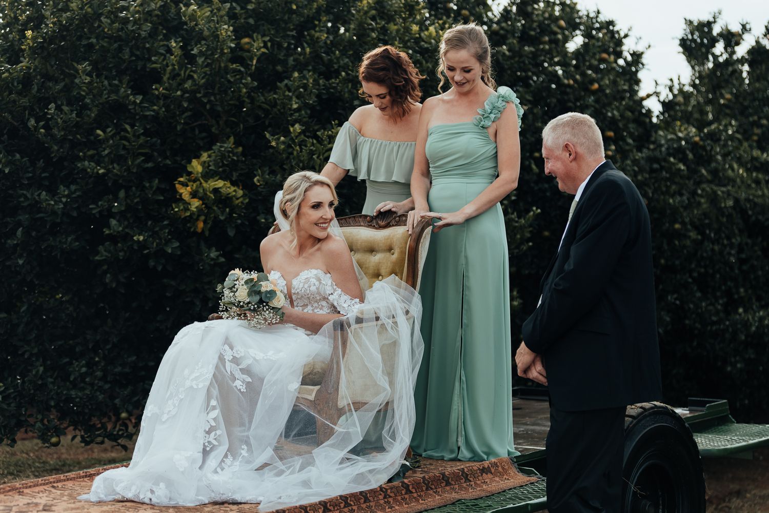 Wedding party members stand on a tractor platform during an outdoor orchard ceremony.