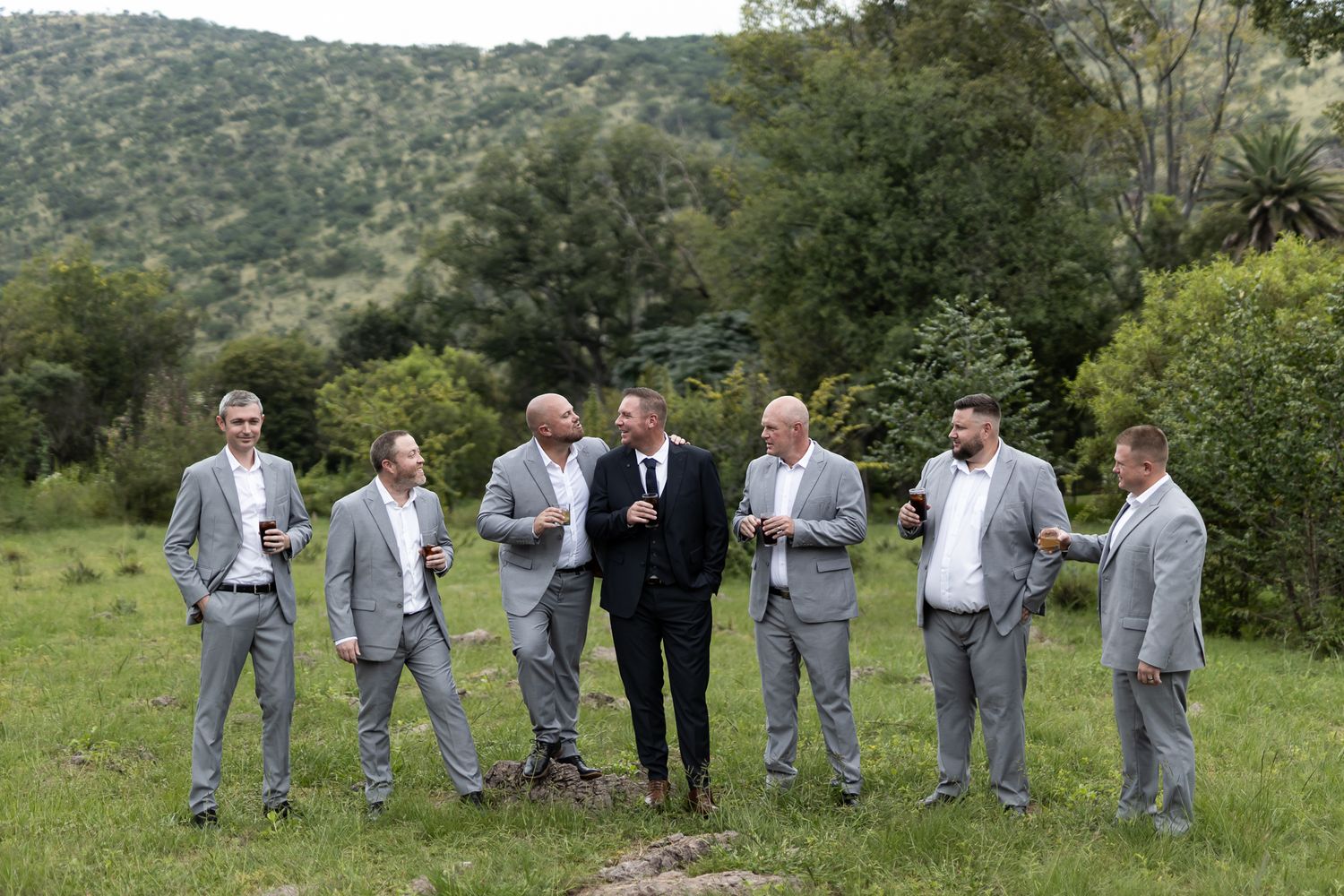 A groom in black suit stands with groomsmen wearing light gray suits in an outdoor rustic setting.