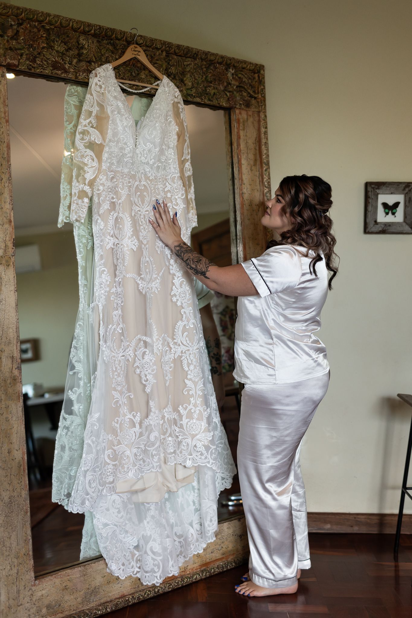 Bride-to-be admiring wedding dress hanging on ornate mirror frame.